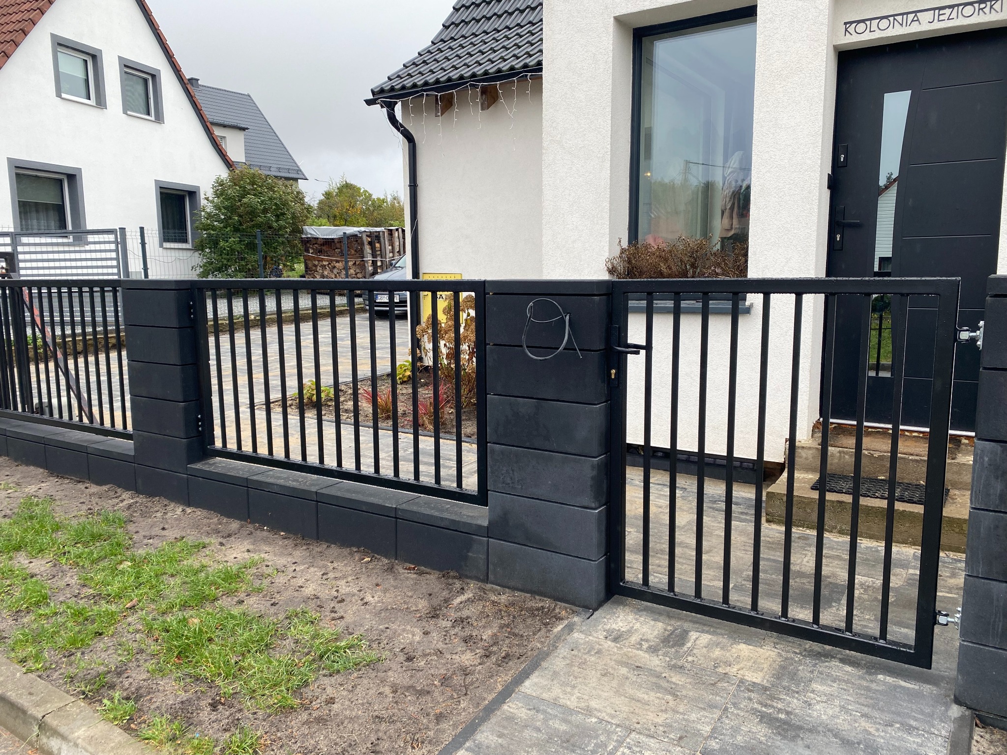 Nowoczesne ogrodzenie metalowe z bramką, czarne słupki z bloczków betonowych, jasny dom w tle, widok z zewnątrz.
