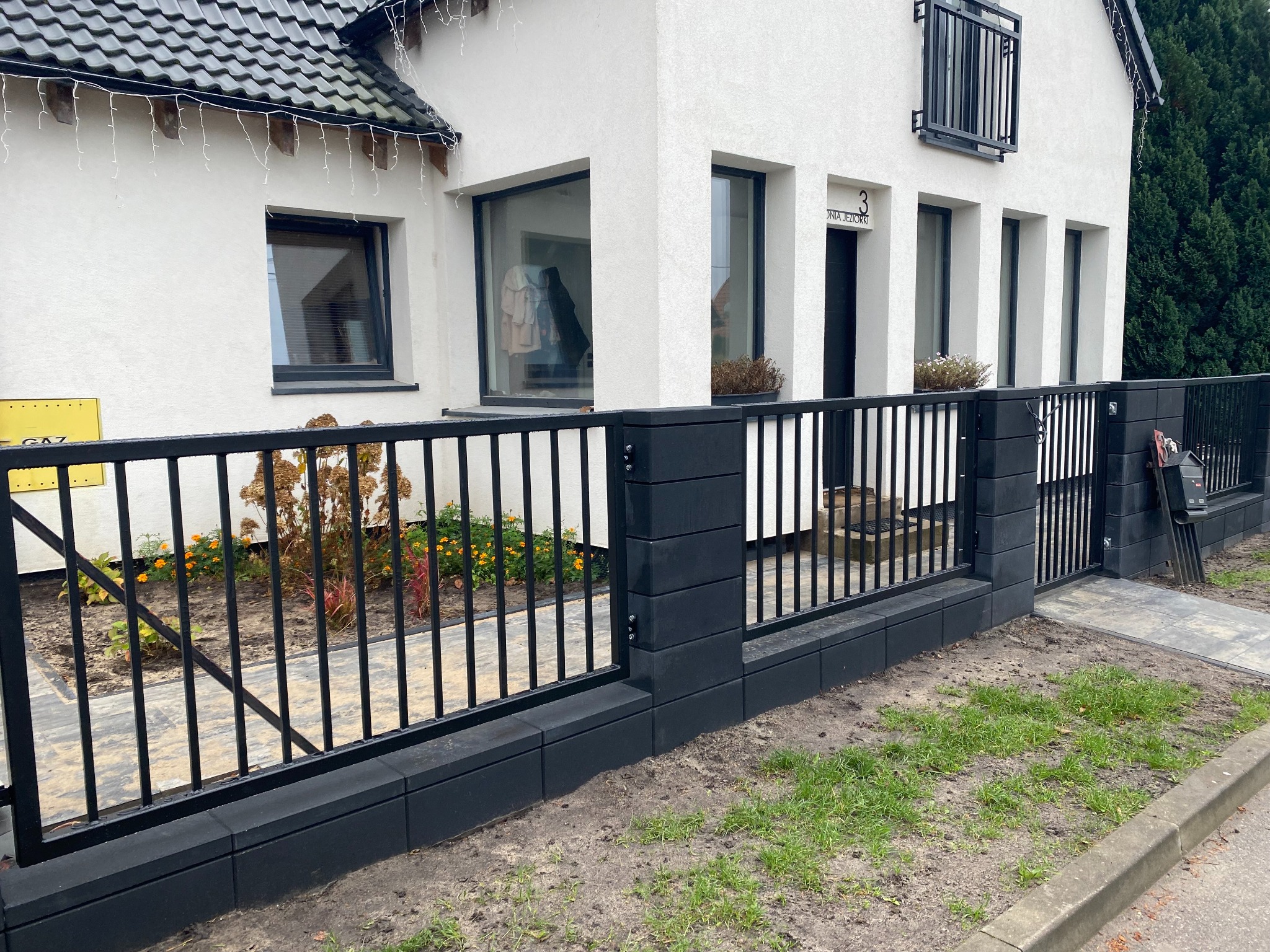 Nowoczesne, czarne ogrodzenie metalowe z furtką i słupkami z bloczków betonowych. Widoczne kwiaty i fragment domu z oknami. Minimalistyczny design.