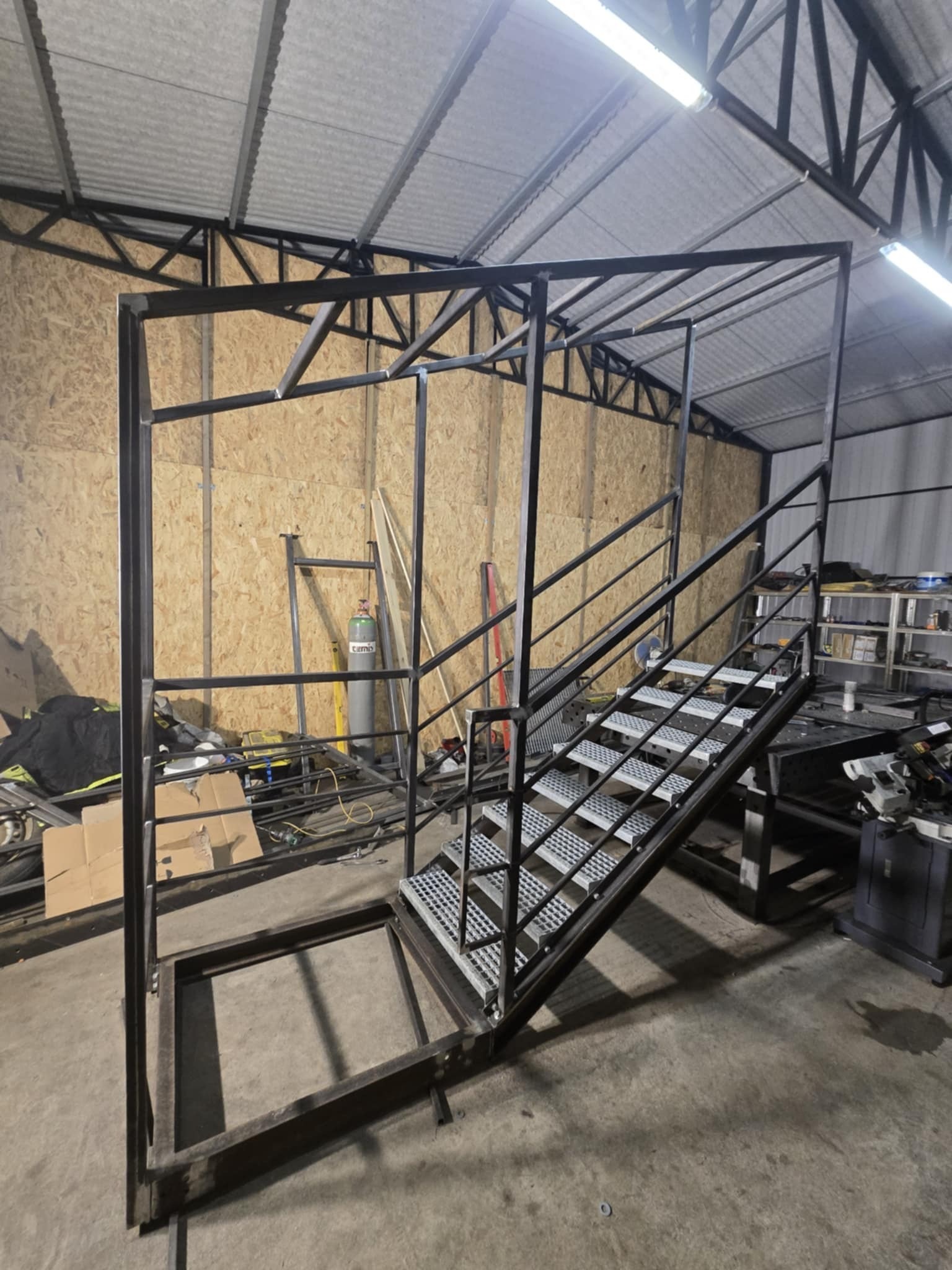Metalowa konstrukcja schodów z balustradą w warsztacie, widok z dołu. Surowy stan, spawane elementy. W tle ściana z płyty OSB i butla z gazem technicznym.