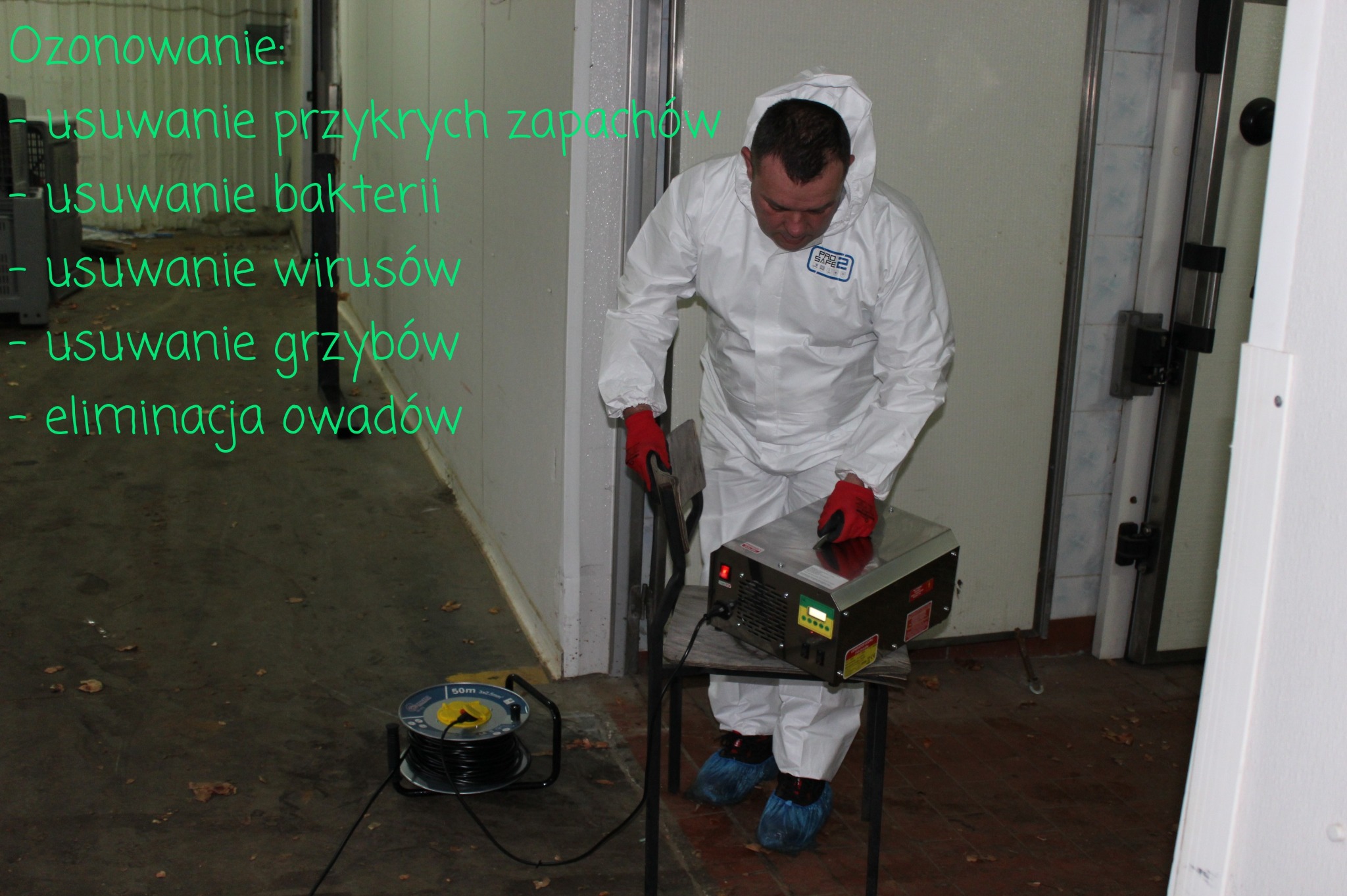 Pracownik w kombinezonie ochronnym i czerwonych rękawicach obsługuje generator ozonu ustawiony na krześle w sterylnym pomieszczeniu, obok widoczny kabel zasilający.