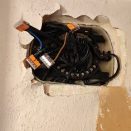 Otwór w ścianie z widocznymi przewodami elektrycznymi połączonymi złączkami WAGO, widoczne fragmenty starej tapety.