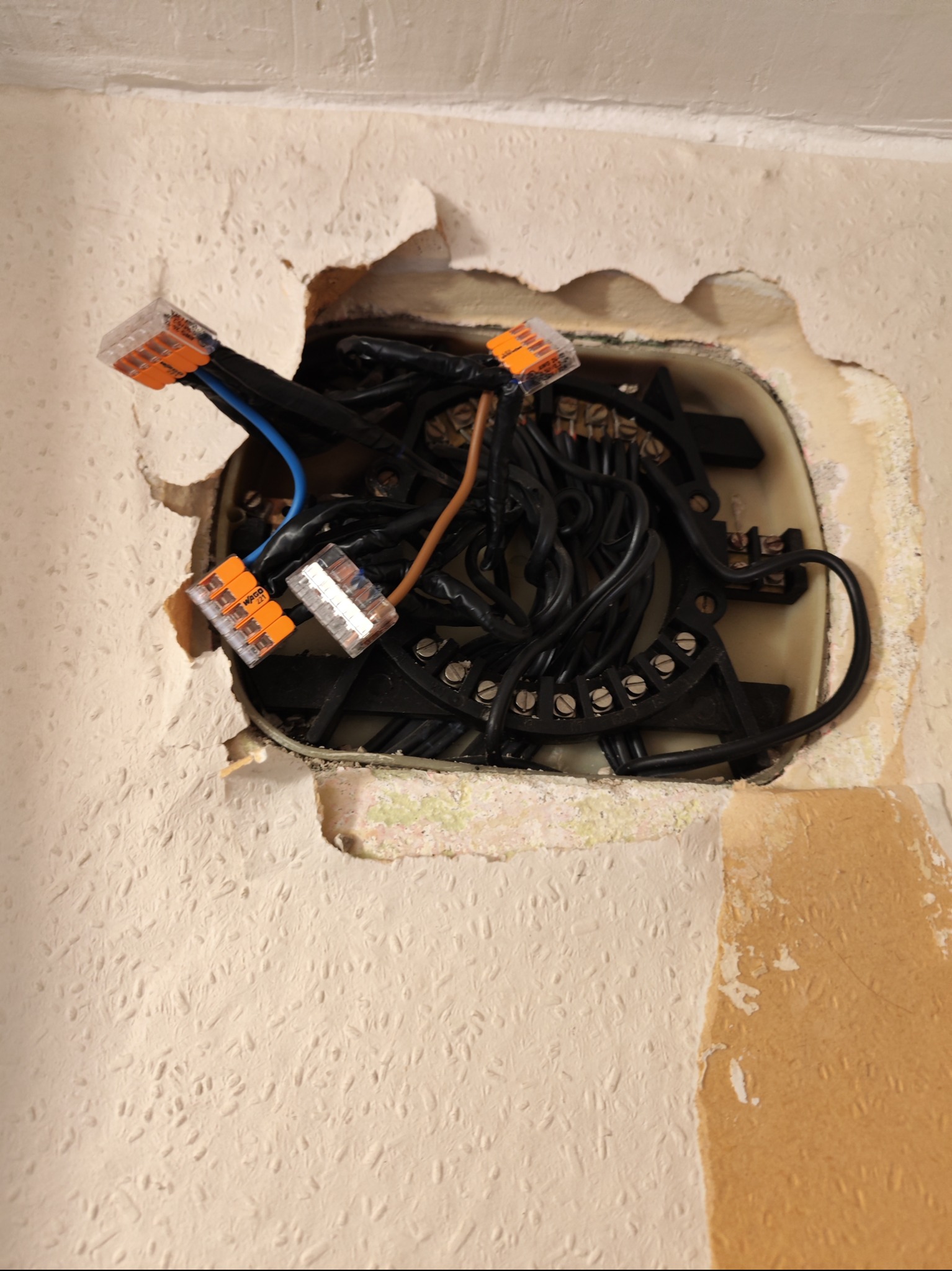 Otwór w ścianie z widocznymi przewodami elektrycznymi połączonymi złączkami WAGO, widoczne fragmenty starej tapety.