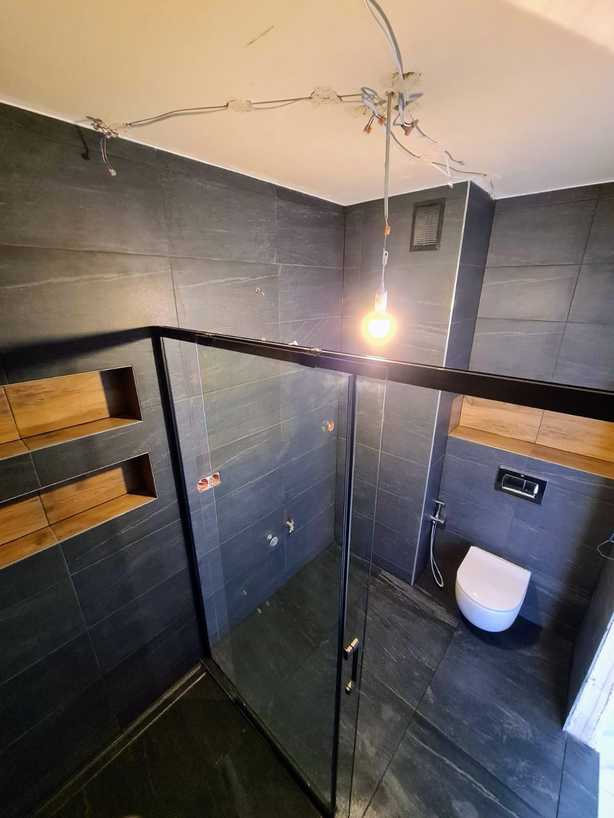 Wykończona łazienka z ciemnymi płytkami, kabiną prysznicową ze szklanymi drzwiami i drewnianymi wnękami. Widoczne niedokończone prace elektryczne na suficie.
