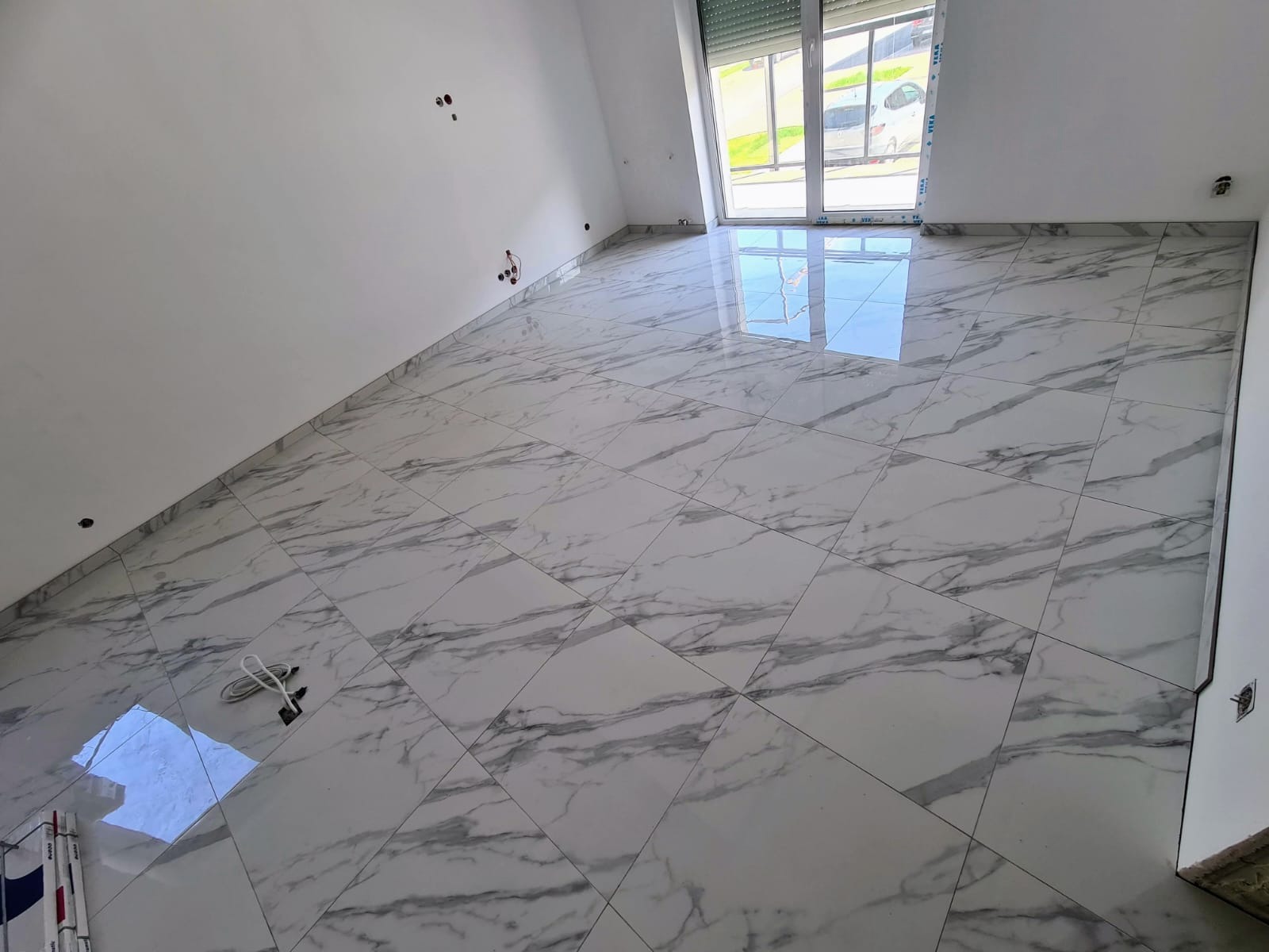 Położone białe płytki imitujące marmur na podłodze w jasnym pokoju z oknem. Widoczne kable wystające ze ściany.