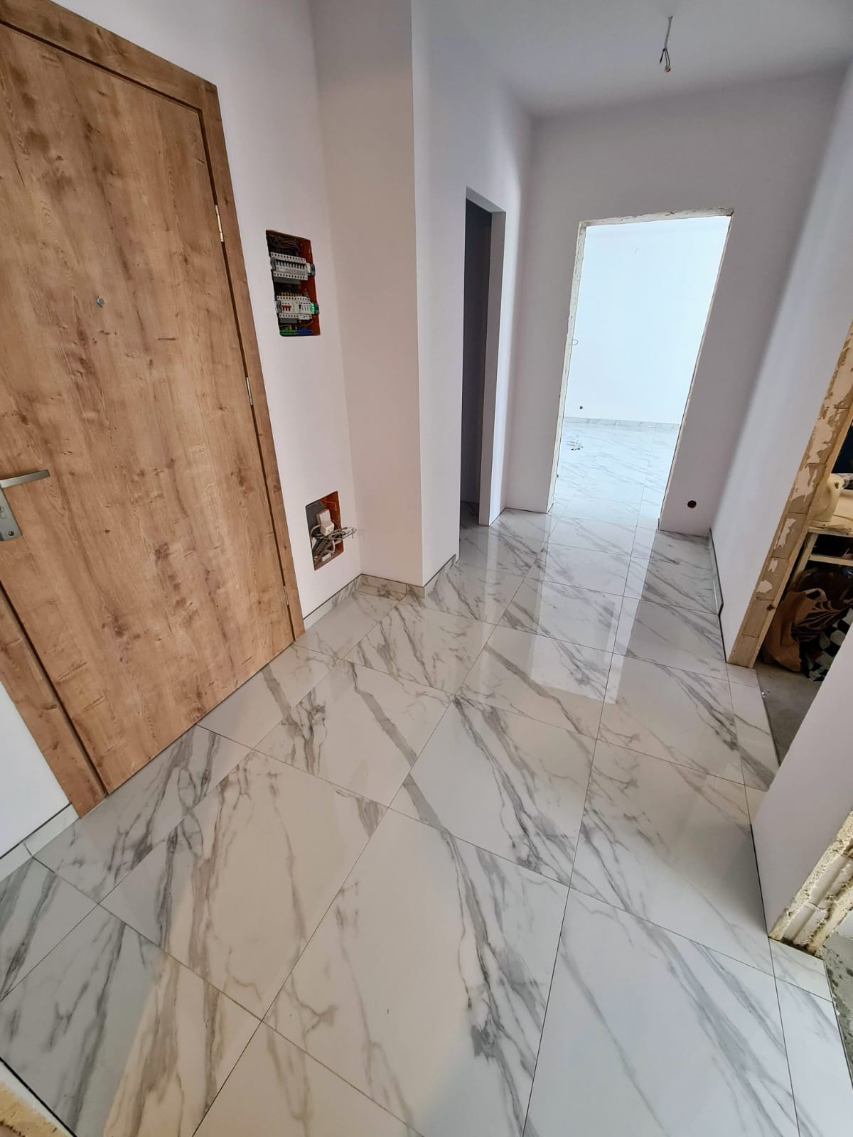 Białe marmurowe płytki podłogowe z szarymi żyłkami w nowym korytarzu. Widoczne drzwi z drewnianą okleiną i rozdzielnia elektryczna na ścianie.