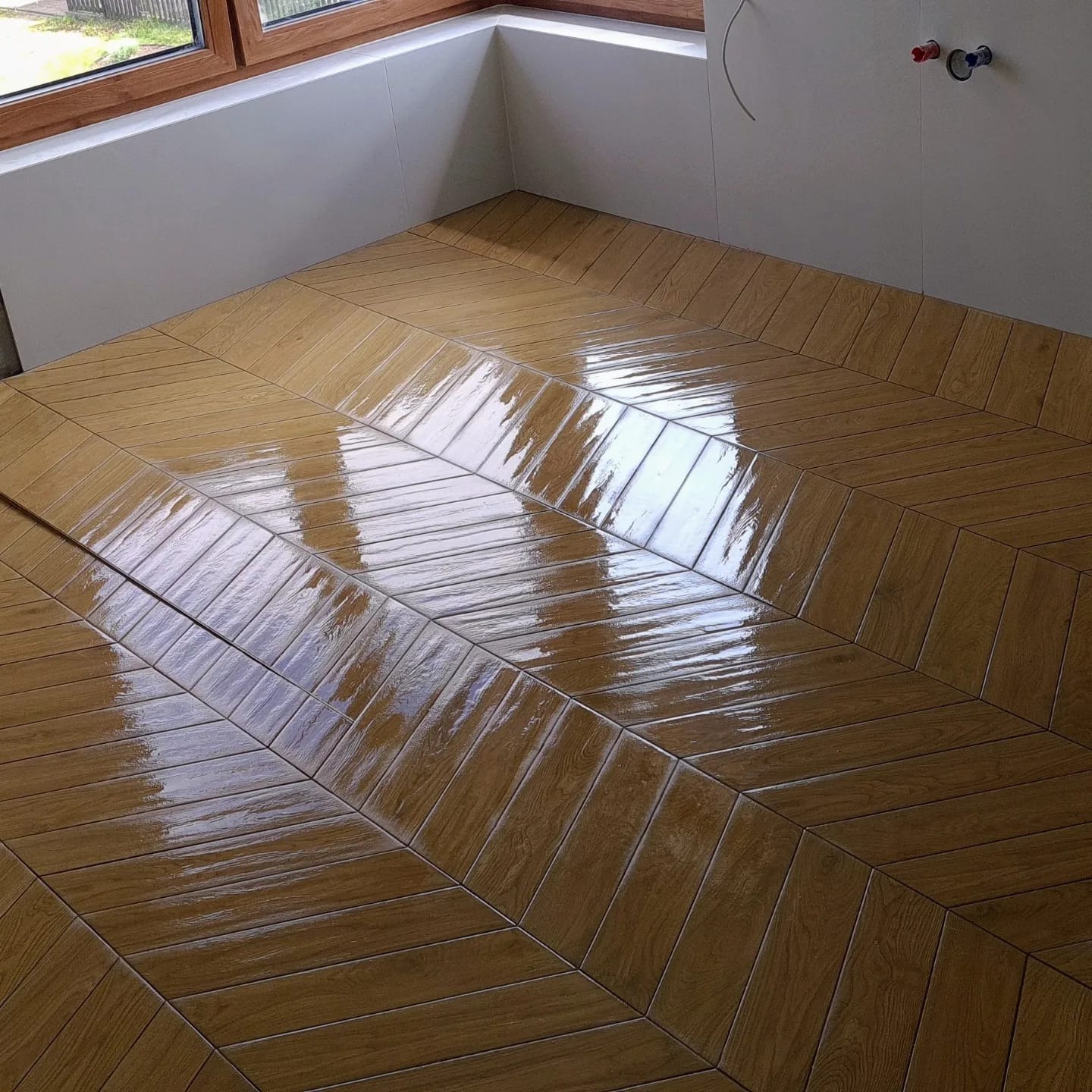 Ukończona podłoga w łazience w jodełkę, z płytek imitujących drewno, Szczecin. Widoczne okno i fragment ściany z instalacją wodną.