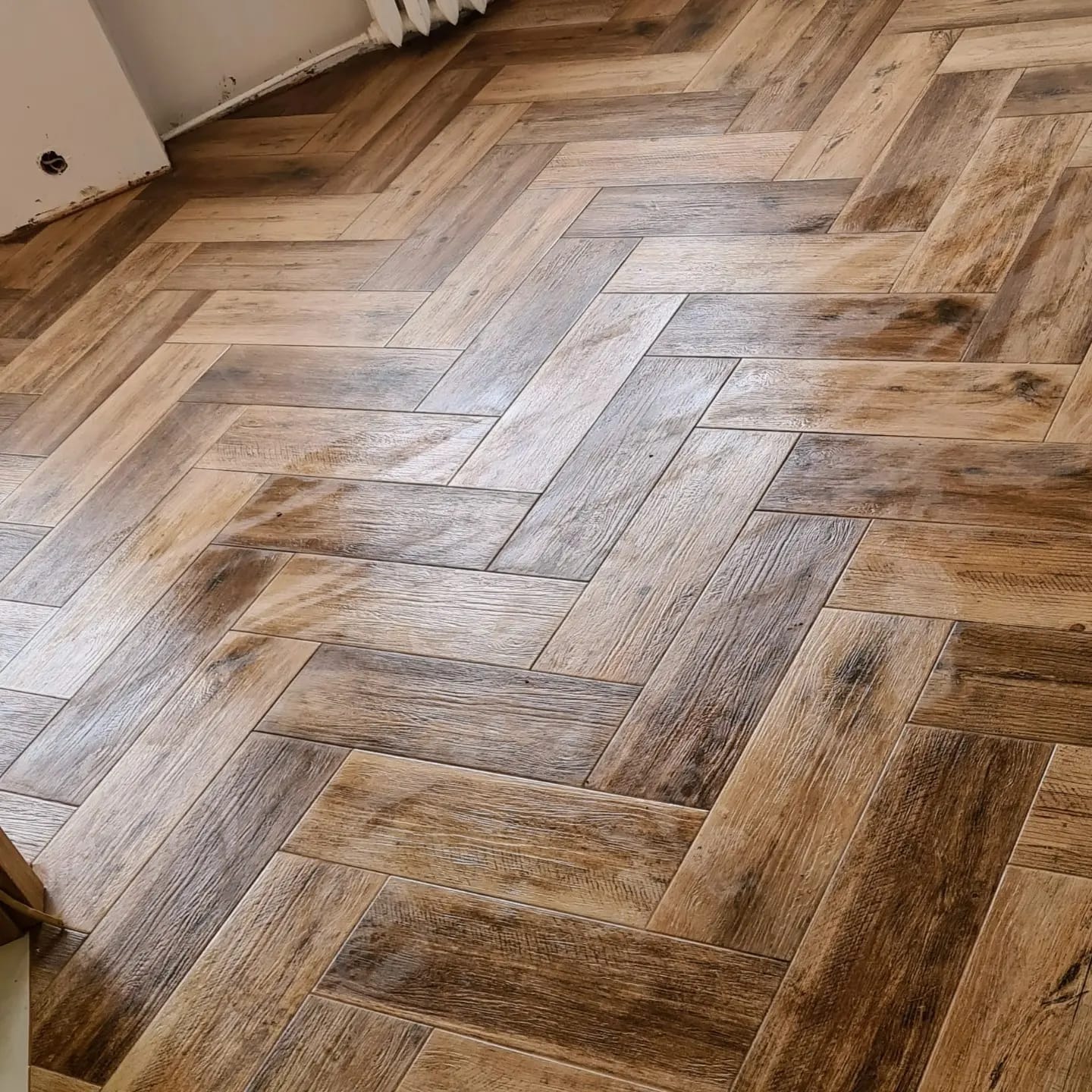 Parkiet ułożony w jodełkę z drewnopodobnych płytek w odcieniach brązu i beżu. Widoczny fragment ściany i grzejnika. Wykończona podłoga.