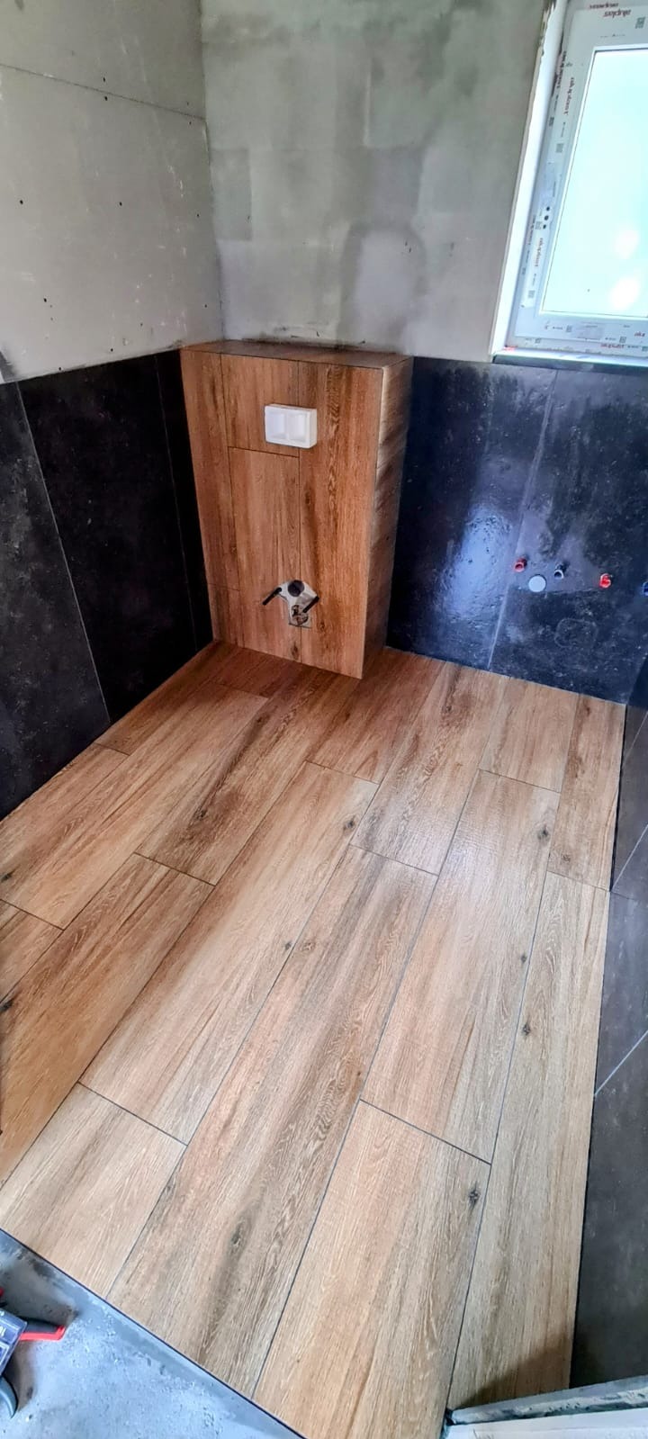 Wykończona łazienka: drewnopodobne płytki na podłodze i czarne na ścianach, zabudowa toalety z drewnopodobnych płytek, okno w tle.