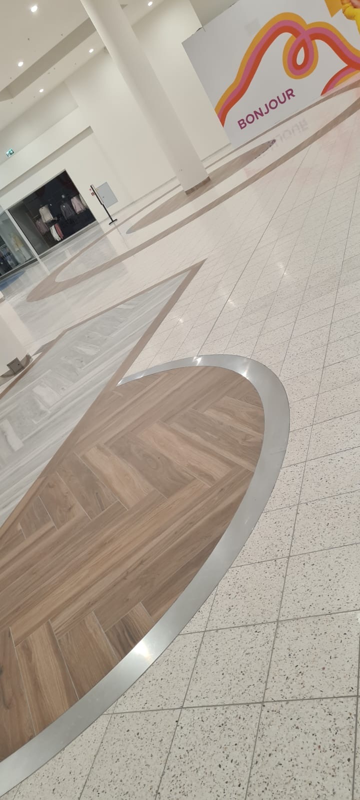 Wzorzysta podłoga w centrum handlowym z połączeniem płytek imitujących drewno w jodełkę i jasnych płytek lastryko, oddzielonych metalową listwą. Widoczny napis 'Bonjour'.
