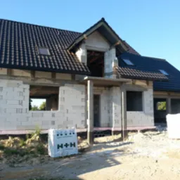 Niedokończony budynek z betonu komórkowego z ciemną dachówką, widoczne pakiety bloczków H+H na placu budowy, okna dachowe zamontowane.