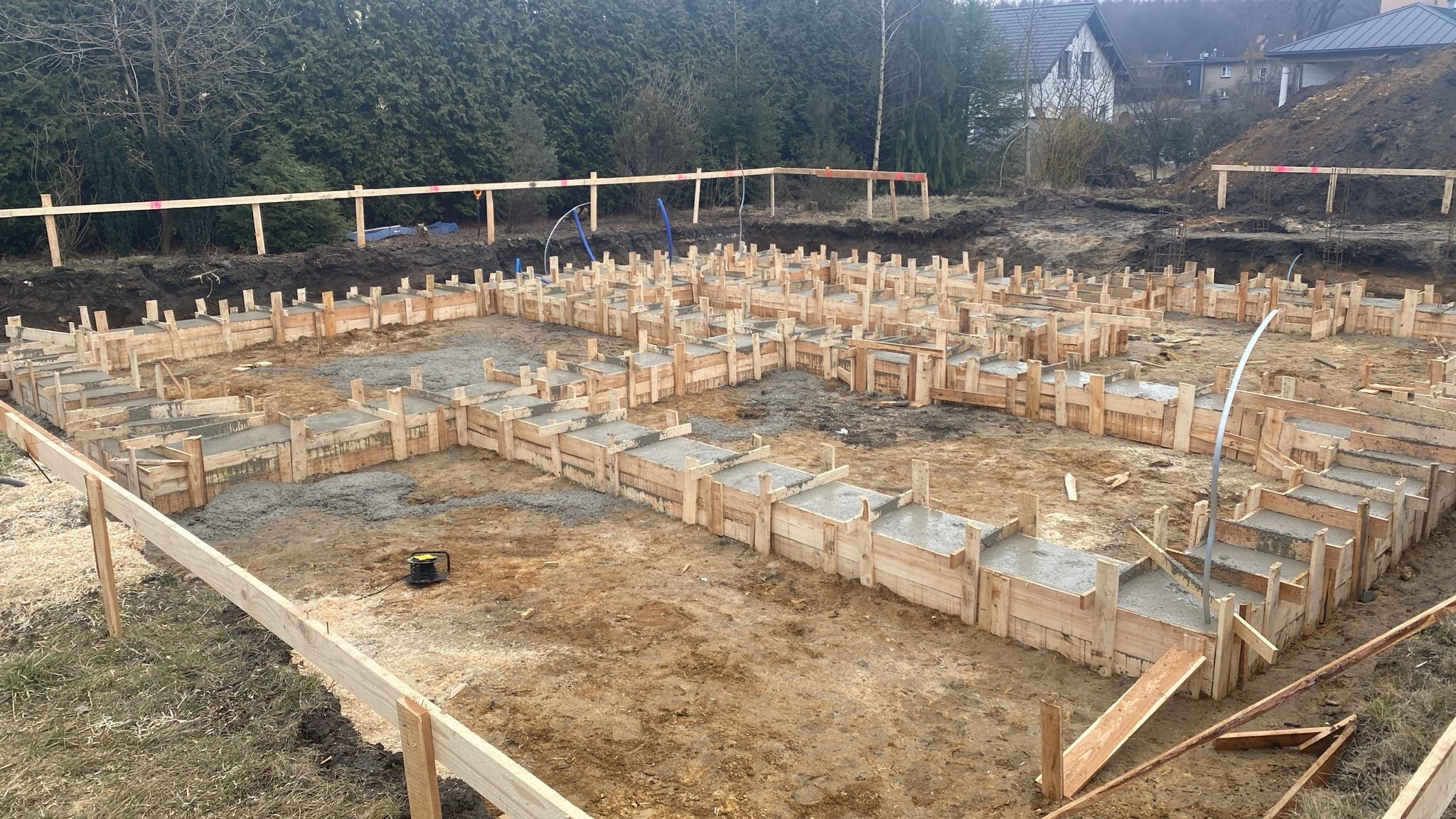 Wylany beton w drewnianych szalunkach tworzących fundamenty domu, widoczne zbrojenia i teren przygotowany pod budowę.