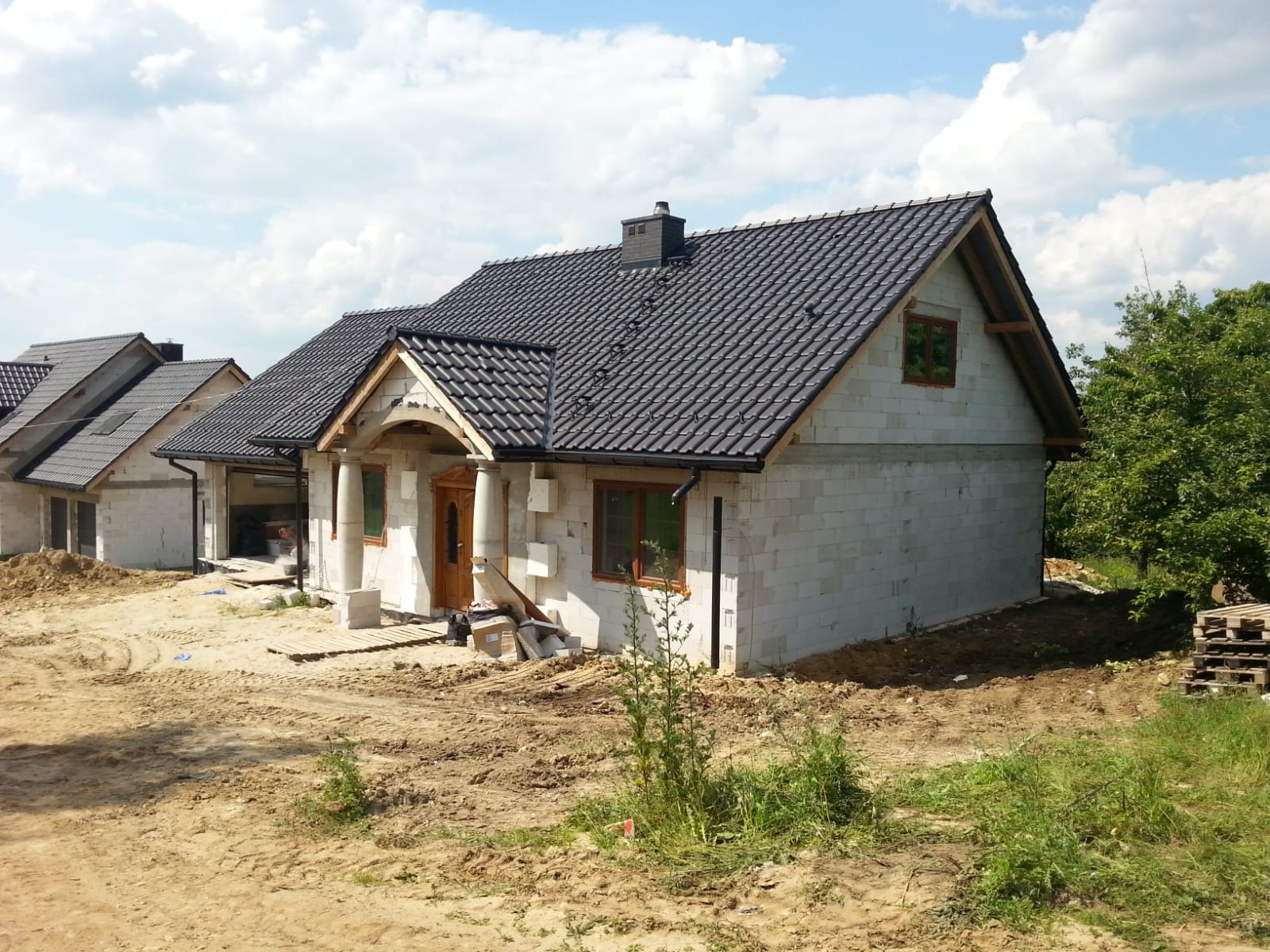 Surowy dom z bloczków betonu komórkowego z ciemną dachówką i drewnianymi oknami, widoczne kolumny i częściowo ułożony chodnik przed wejściem, ziemia wokół domu. W tle kolejne domy w stanie surowym.