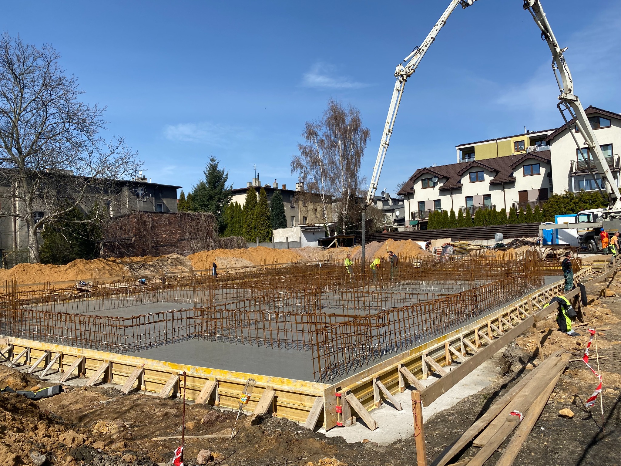 Wylewanie betonu na przygotowany zbrojony fundament z drewnianym szalunkiem, widoczny wysięgnik pompy do betonu i pracownicy w odblaskowych kamizelkach.