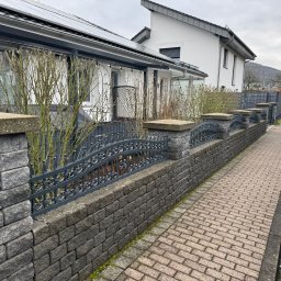 Arkadiusz kaczanowski - Niski murek z szarej kostki betonowej zwieńczony metalowym, kutym ogrodzeniem z dekoracyjnymi łukami, oddzielający chodnik od posesji z białym domem i zielenią.