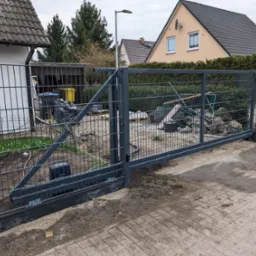 Szara, przesuwna brama panelowa z automatyką, widok na posesję z gruzem i workami, w tle domy jednorodzinne.