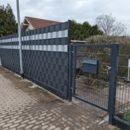 Nowoczesne ogrodzenie panelowe w kolorze grafitowym z poziomymi wstawkami w odcieniach szarości, przylegające do furtki z metalową skrzynką na listy, na tle brukowanego chodnika i zabudowy...