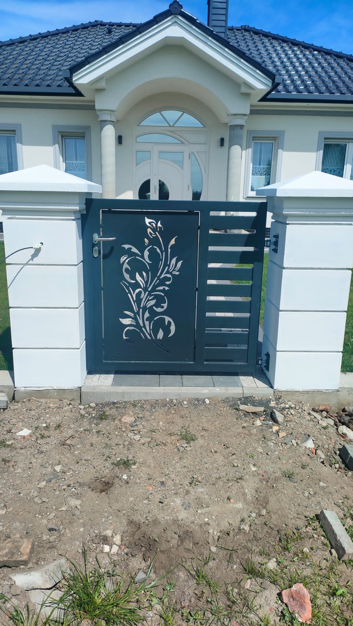 Nowoczesna, grafitowa furtka z laserowo wycinanym motywem roślinnym, osadzona między białymi, kwadratowymi słupkami z betonu architektonicznego, na tle domu z ciemną dachówką.
