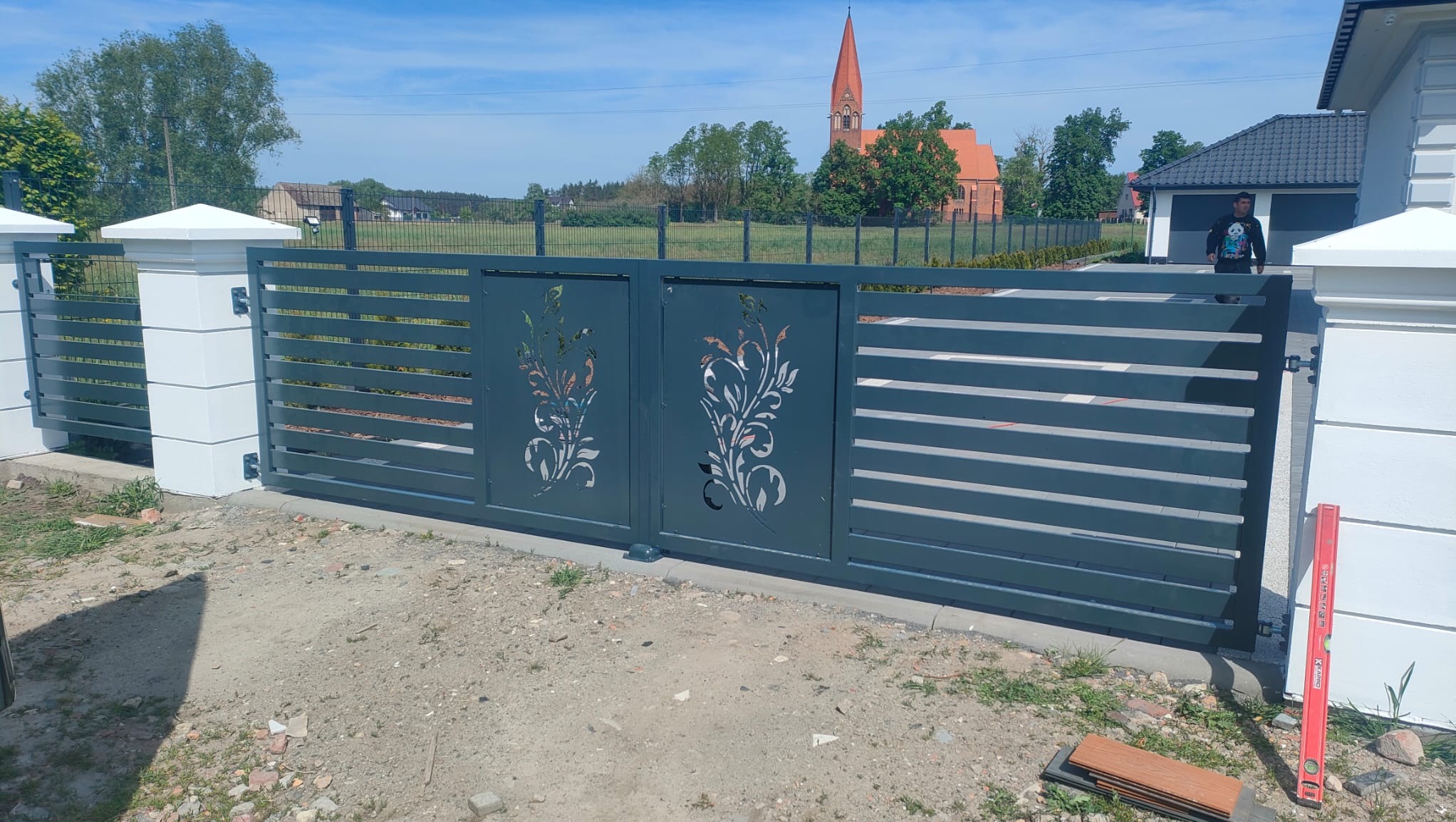 Przesuwna, szara brama z poziomymi szczeblami i wycinanym laserowo motywem roślinnym, zamontowana między białymi słupkami. W tle widoczny kościół z czerwoną wieżą i osoba w czarnej bluzie.