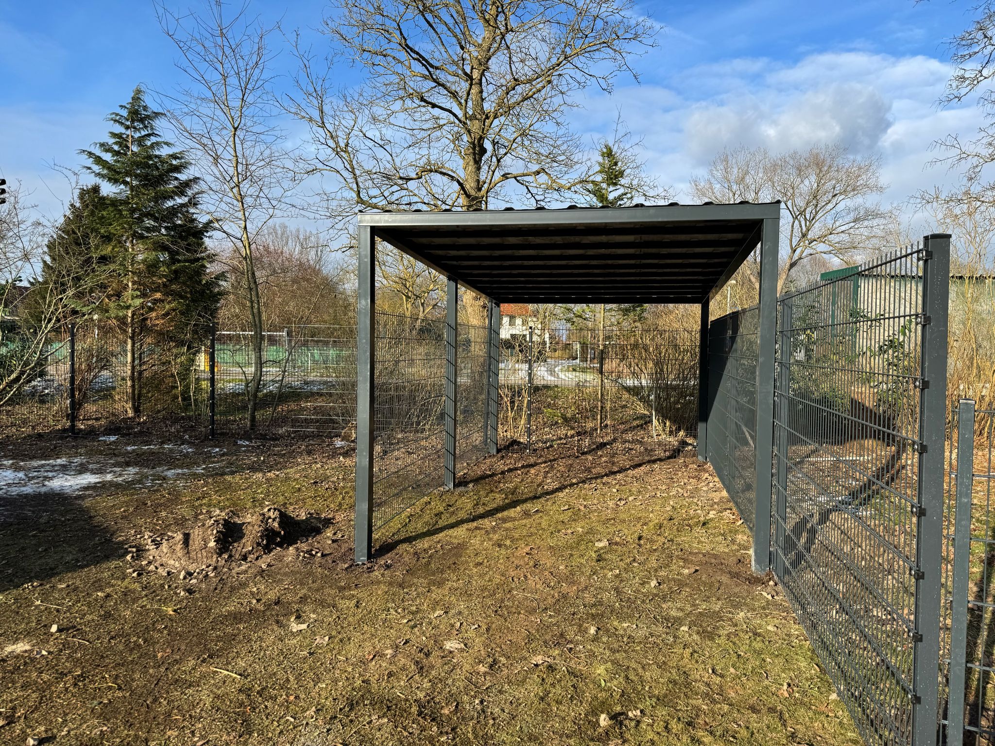 Szara, metalowa konstrukcja z zadaszeniem, prawdopodobnie wiata lub carport, otoczona metalowym ogrodzeniem panelowym na tle ogrodu z drzewami i błękitnym niebem.