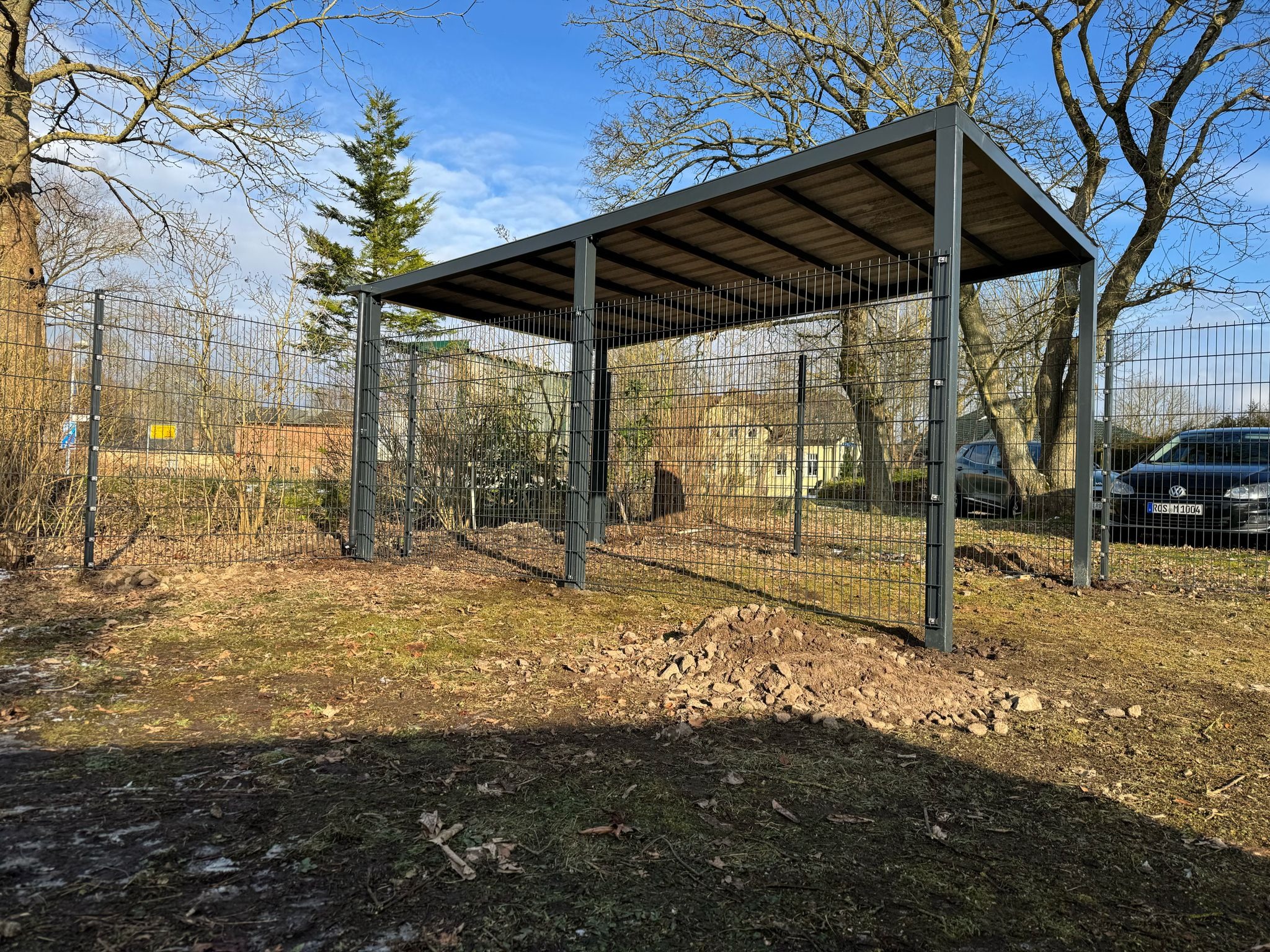 Nowoczesna, szara konstrukcja zadaszenia z siatkową obudową, otoczona metalowym ogrodzeniem panelowym na nierównym terenie z widocznymi samochodami w tle.