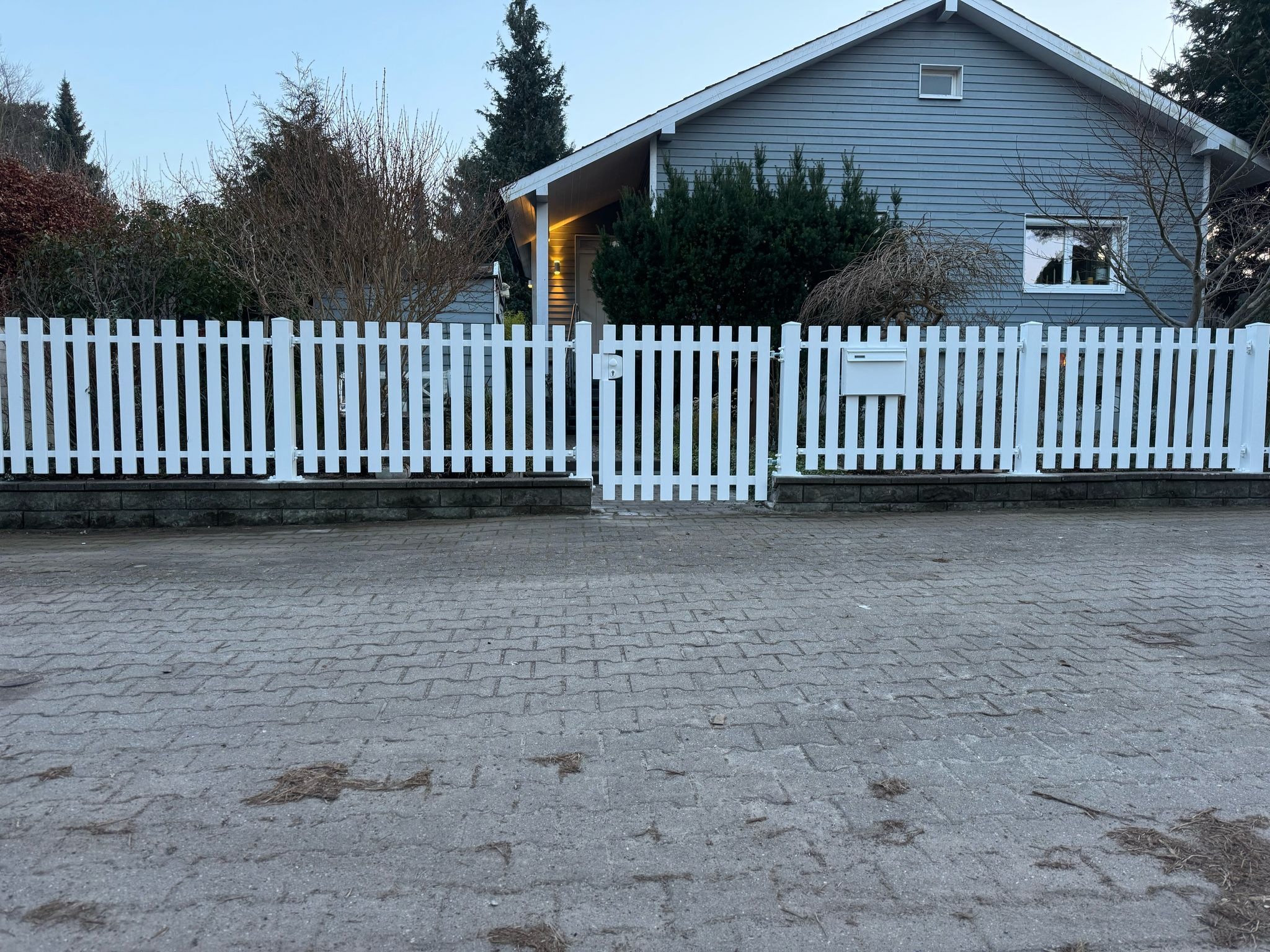 Białe sztachetowe ogrodzenie z furtką przed szarym domem jednorodzinnym z widoczną skrzynką na listy, ujęcie z poziomu chodnika.