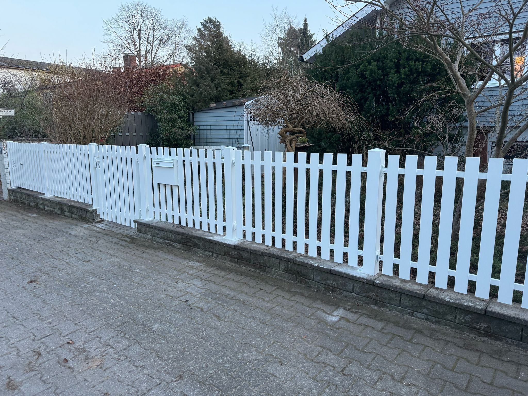 Białe, sztachetowe ogrodzenie z furtką i skrzynką na listy, osadzone na niskim murku z szarej kostki. Tło stanowią domy i drzewa bez liści.