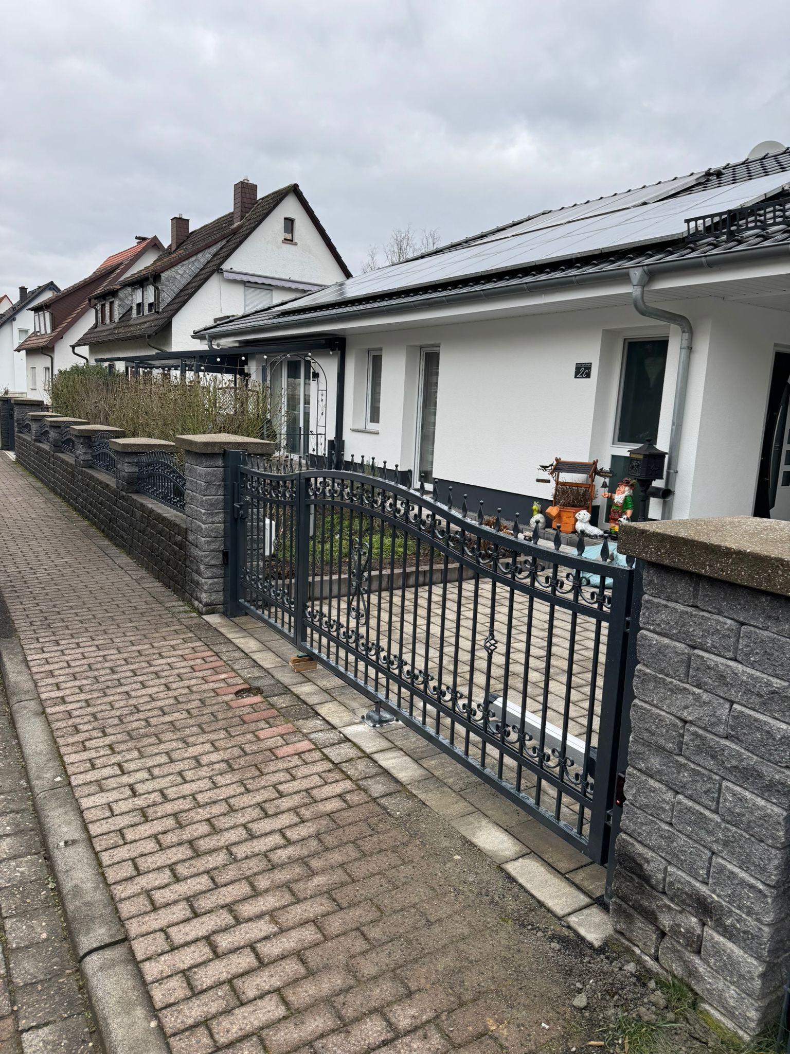 Szara brama wjazdowa z metalowymi zdobieniami i murowanymi słupkami z szarego kamienia, na posesji z białym domem i panelami słonecznymi na dachu.