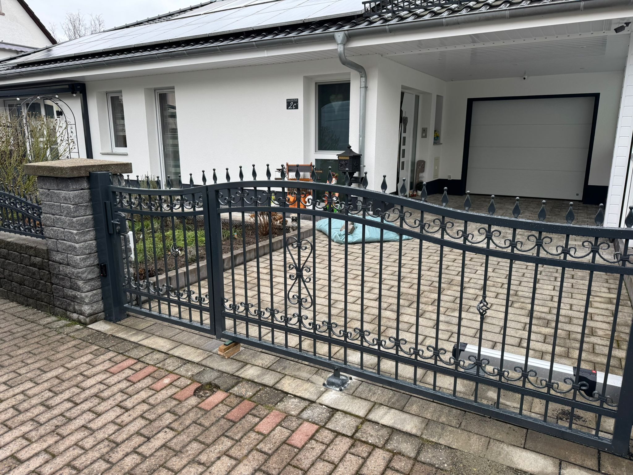 Czarna, metalowa brama wjazdowa z dekoracyjnymi elementami, zamontowana przed nowoczesnym, białym domem z panelami słonecznymi na dachu i brukowanym podjazdem.