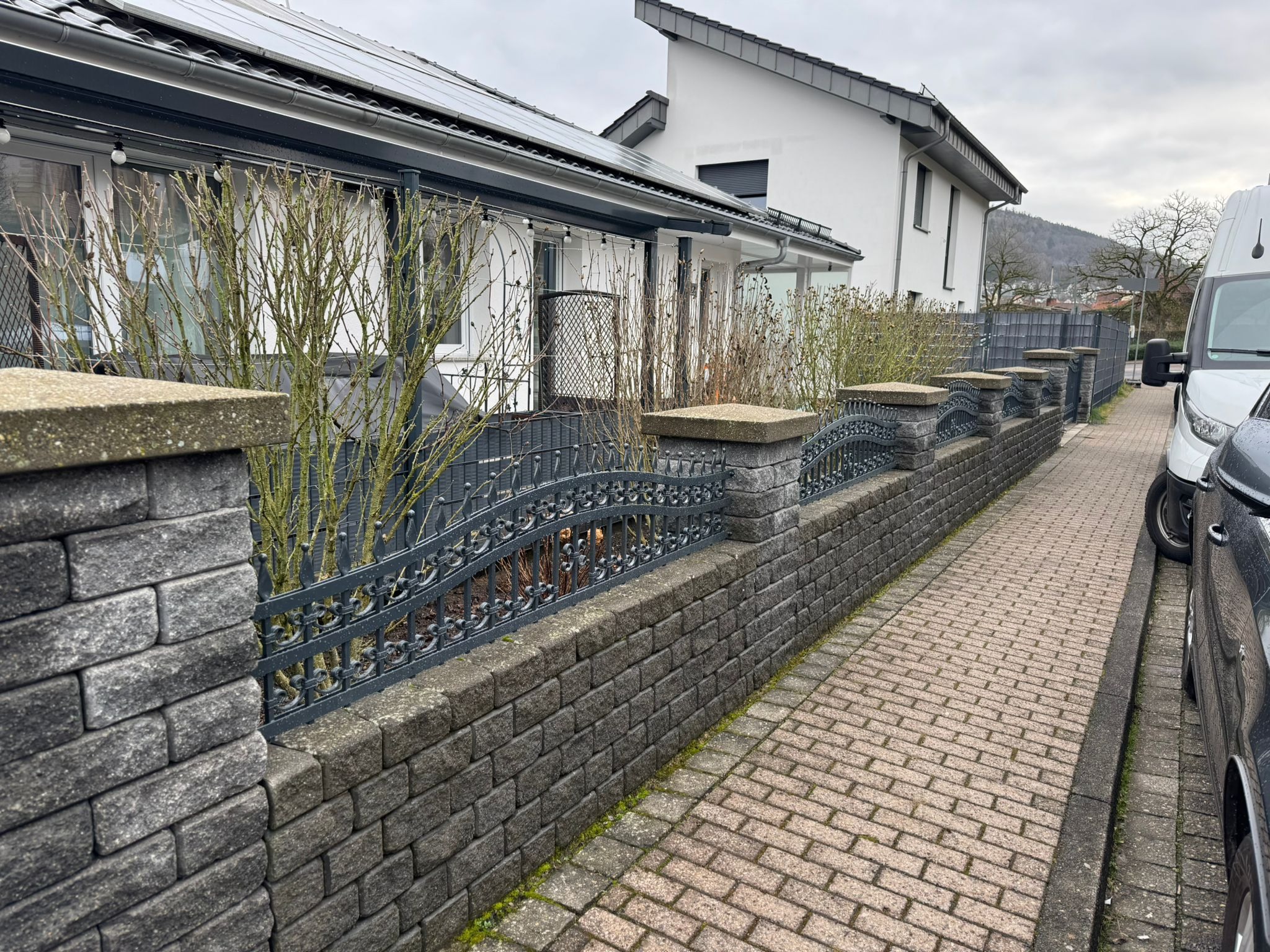 Niski murek z szarej kostki betonowej zwieńczony metalowym, kutym ogrodzeniem z dekoracyjnymi łukami, oddzielający chodnik od posesji z białym domem i zielenią.