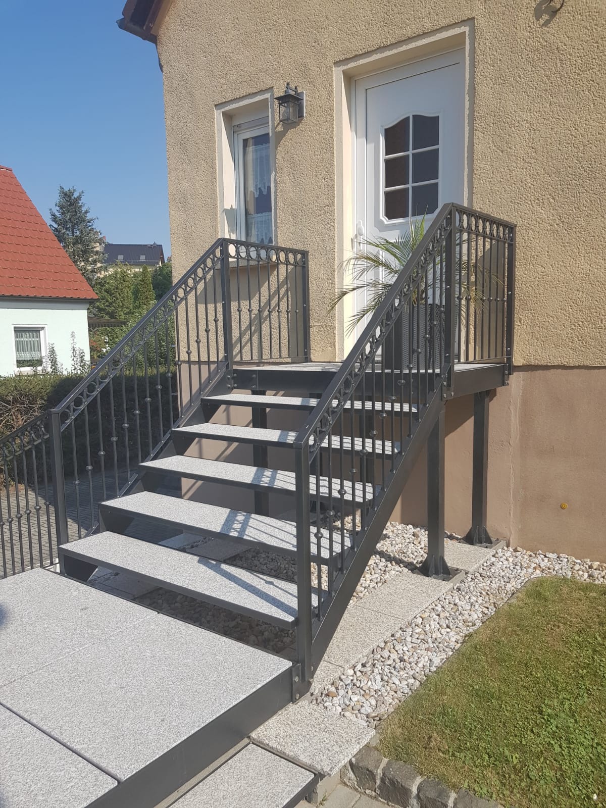 Metalowe schody zewnętrzne z szarymi stopniami prowadzące do drzwi wejściowych, z ozdobną balustradą z motywem kulek, na tle beżowej elewacji domu i zielonego trawnika.