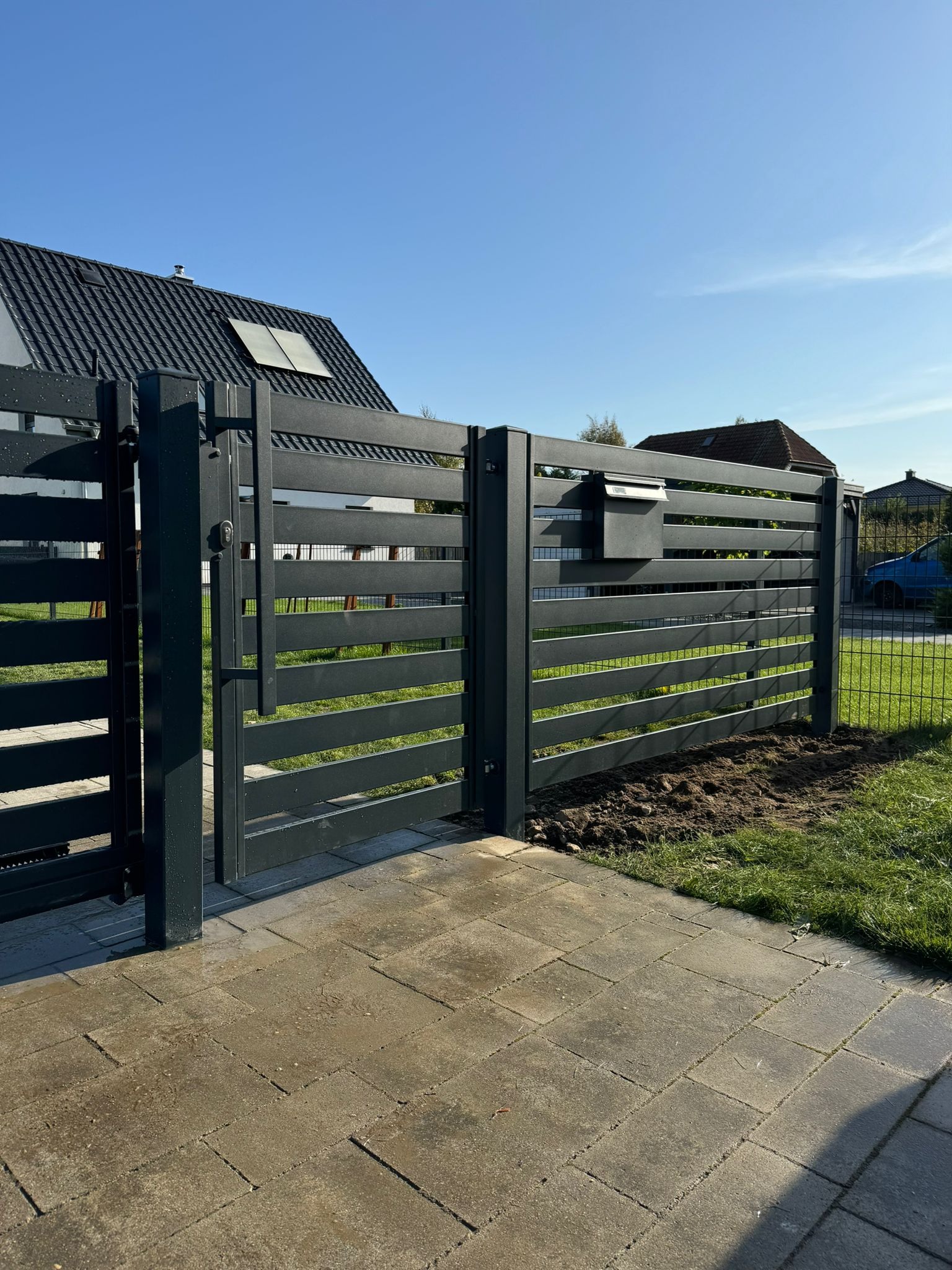 Nowoczesna, pozioma brama wjazdowa z metalu w kolorze antracytowym z wbudowaną skrzynką na listy, widok z zewnątrz na kostkę brukową.