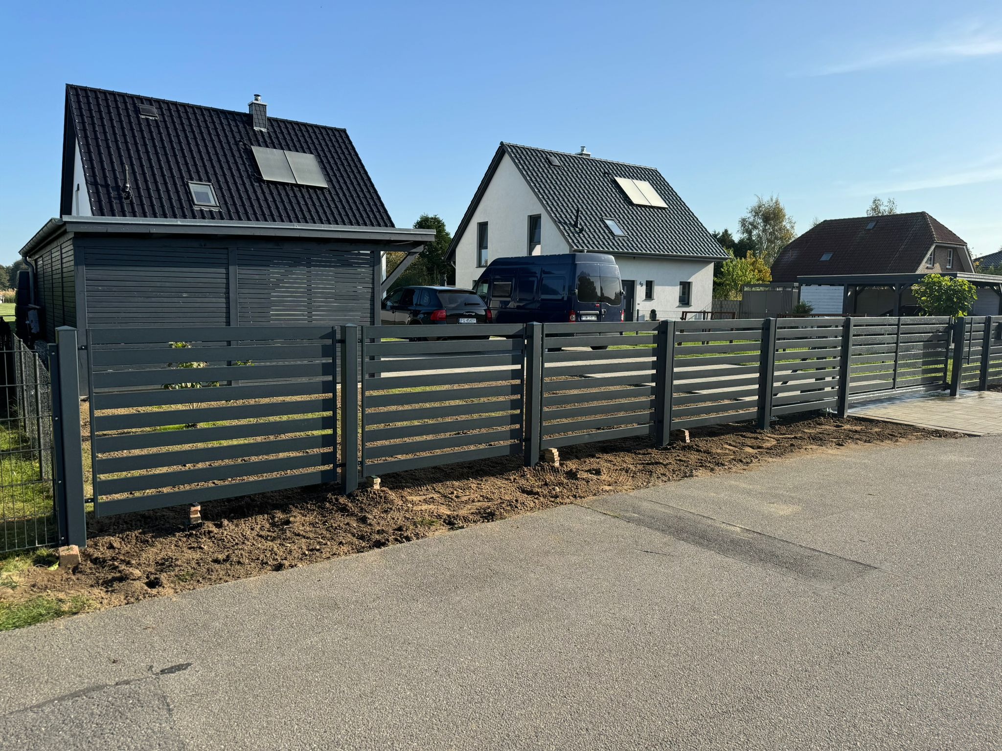 Nowoczesne, poziome przęsła w kolorze grafitowym tworzą ogrodzenie posesji z widocznymi dwoma domami w tle, świeżo obsypane ziemią u podstawy.