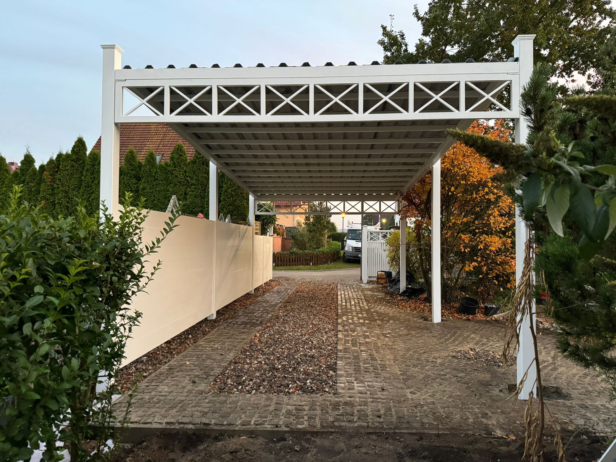 Nowoczesna, biała konstrukcja zadaszenia wjazdu z dekoracyjnymi krzyżami, widok z zewnątrz na brukowaną drogę z kamiennym pasem pośrodku, obok białe ogrodzenie panelowe i zieleń