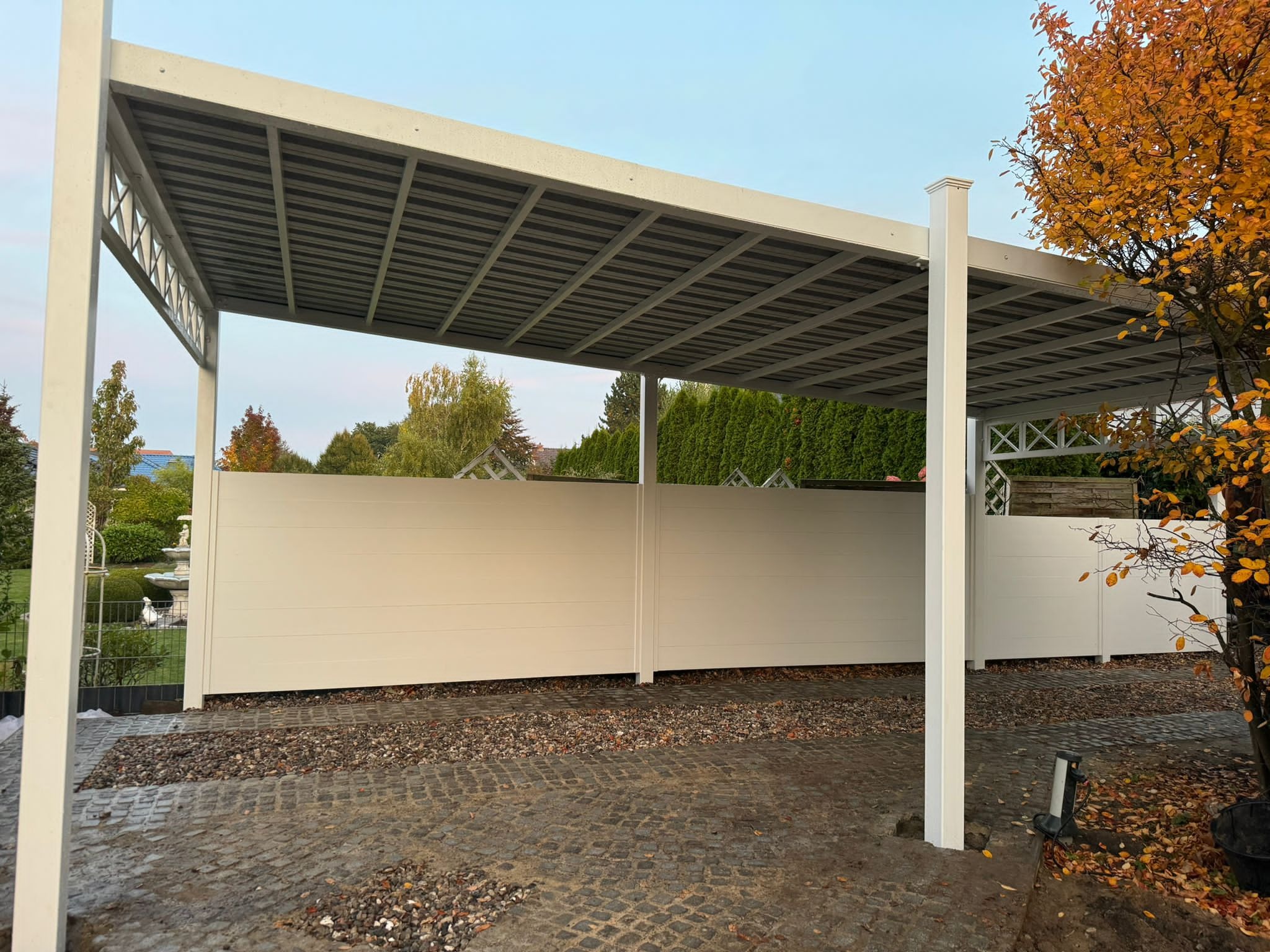 Nowoczesna wiata garażowa z białymi metalowymi słupami i płotem, widok z zewnątrz na brukowanym podjeździe z drzewem o jesiennych liściach.