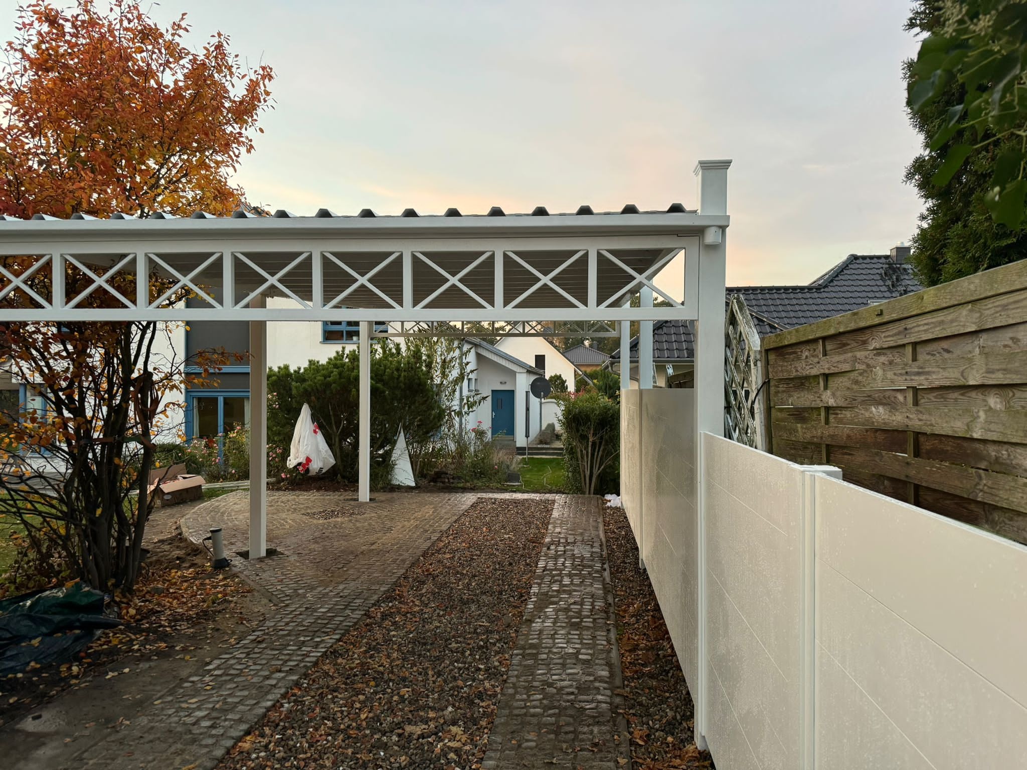 Nowoczesna, biała wiata garażowa z kratownicą i słupkami, zintegrowana z panelowym ogrodzeniem w kolorze złamanej bieli, widok na brukowany podjazd z opadłymi liśćmi.