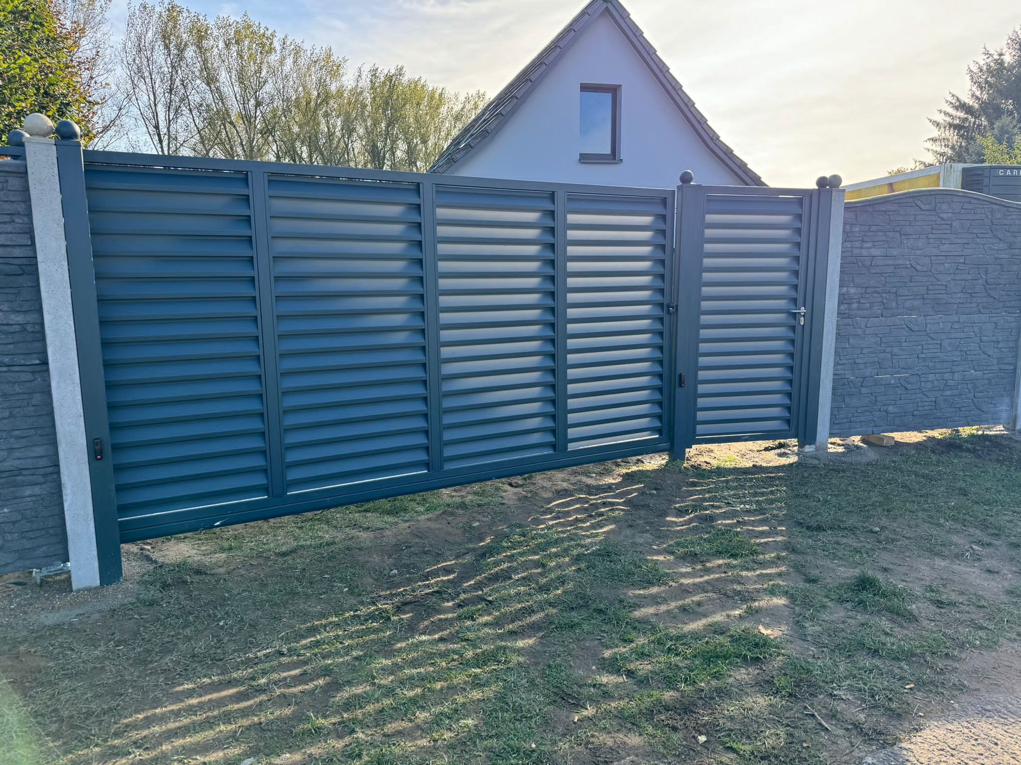 Nowoczesna przesuwna brama wjazdowa z poziomych profili w kolorze grafitowym, zamontowana na betonowych słupkach z dekoracyjnymi kulami, widok na dom z szarym dachem i trawnik z cieniami.