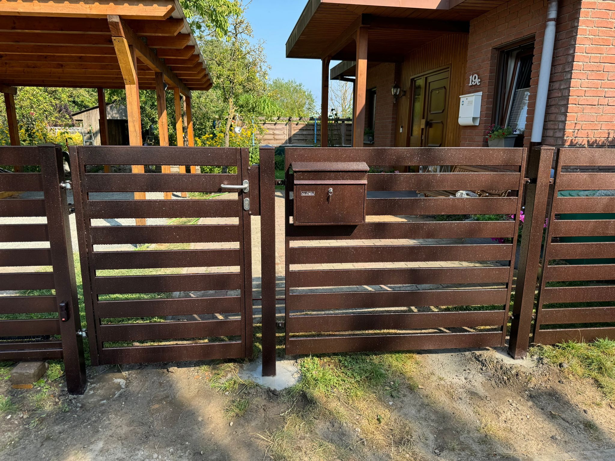 Nowoczesna, pozioma brama wjazdowa z metalu w kolorze brązowym, z zamontowaną skrzynką na listy, na tle domu z czerwonej cegły i zieleni ogrodu.