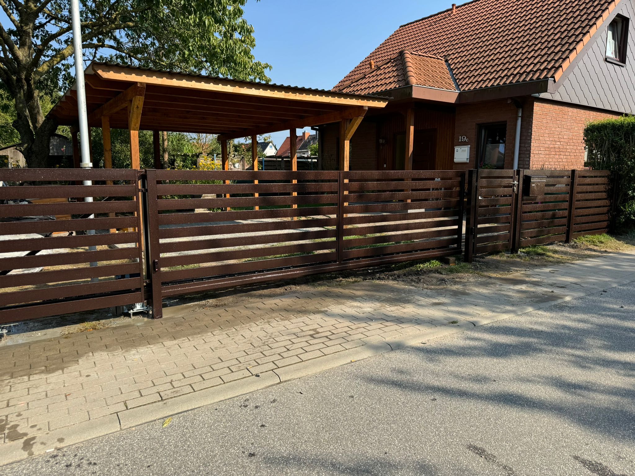 Nowoczesna, brązowa brama przesuwna z poziomych profili, z widocznym mechanizmem automatycznego otwierania, na tle domu z drewnianą wiatą.