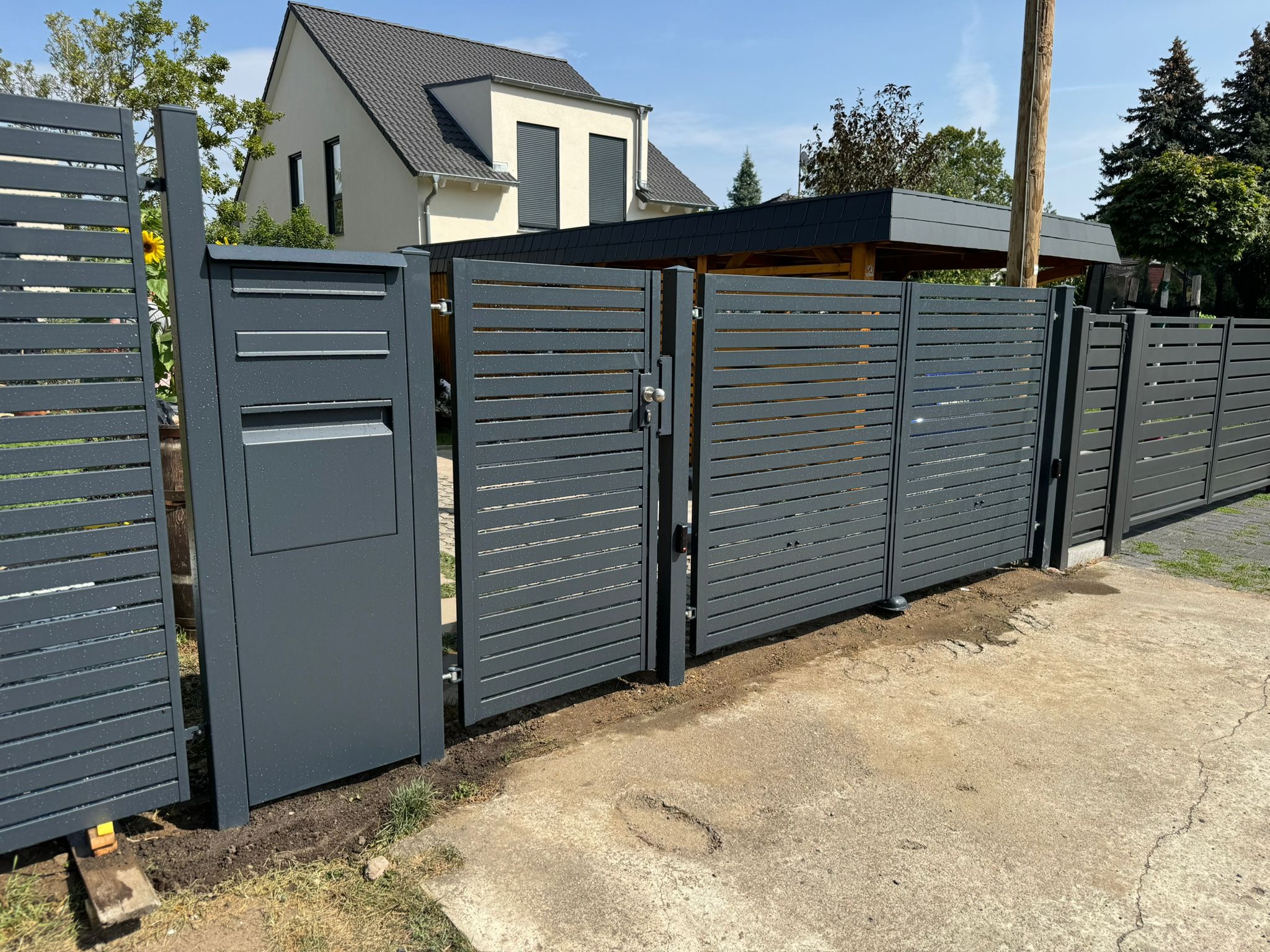 Nowoczesna, pozioma, szara konstrukcja z przesuwną bramą i skrzynką pocztową, widok z zewnątrz na tle domu z szarą dachówką i zielenią.