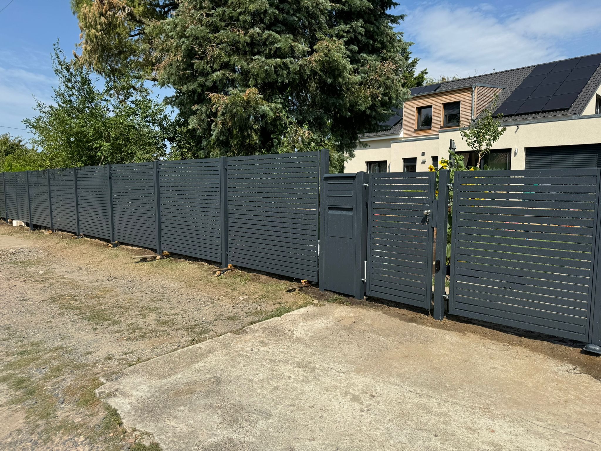 Nowoczesne, poziome, szare panele tworzące ogrodzenie z furtką i skrzynką na listy, na tle domu z panelami słonecznymi.