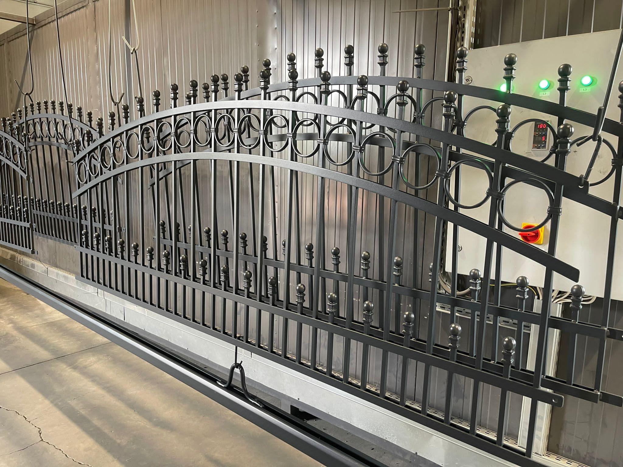 Nowoczesna, przesuwna brama wjazdowa z metalu, z dekoracyjnymi okręgami i kulkami, w trakcie procesu malowania proszkowego w komorze lakierniczej, z widocznym panelem sterowania.