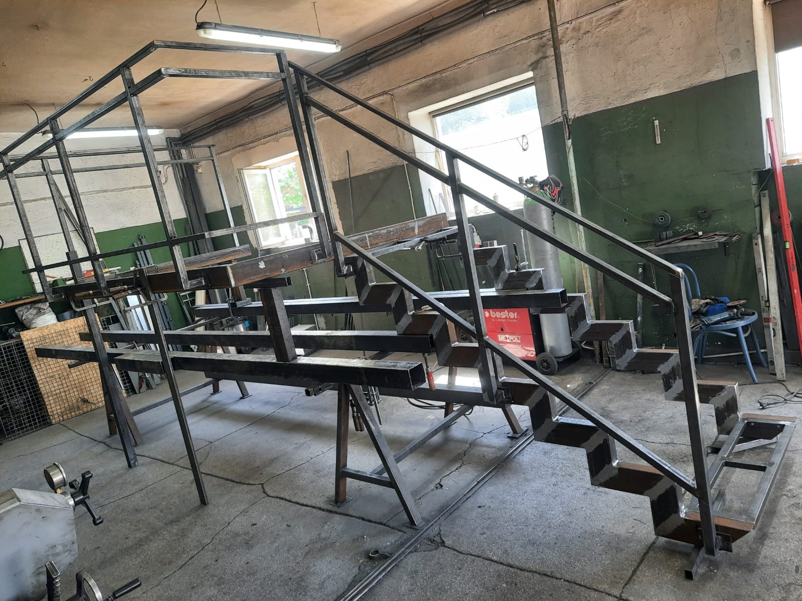Surowa, metalowa konstrukcja schodów w trakcie spawania w warsztacie, widoczna spawarka Bester i okno w tle.