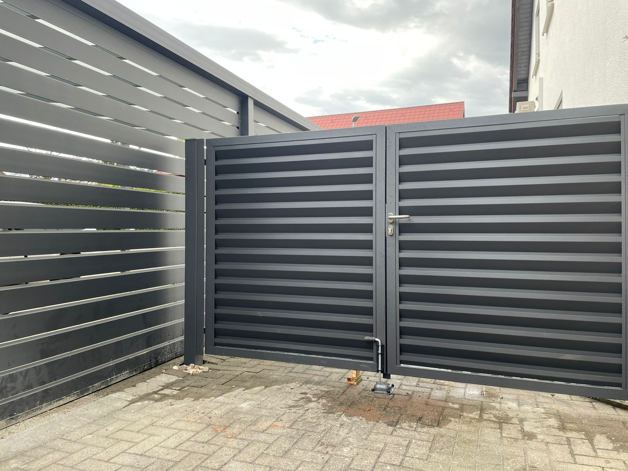 Nowoczesna, pozioma, metalowa brama wjazdowa w kolorze grafitowym, z widocznym mechanizmem otwierania i częścią ogrodzenia w tle, na tle kostki brukowej i budynku.