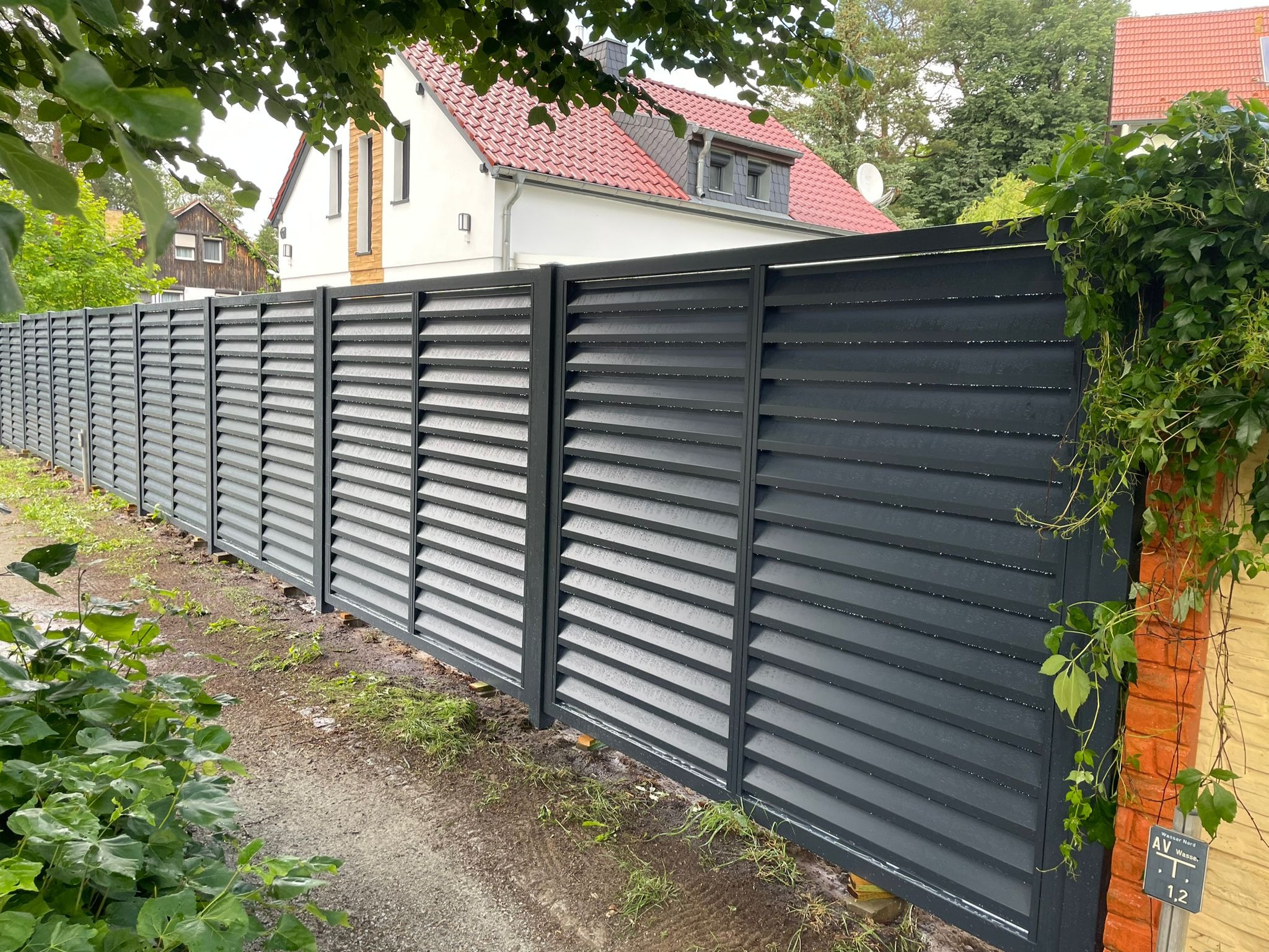 Nowoczesne, poziome przęsła w kolorze grafitowym tworzą ogrodzenie posesji z białym domem w tle, z elementami zieleni pnącej się po ceglanym słupku.