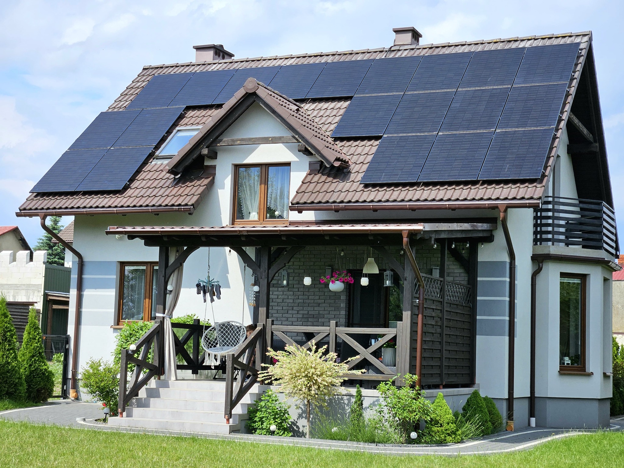 Dom jednorodzinny z brązowym dachem i panelami słonecznymi na połaci dachu. Widoczny zadaszony taras z huśtawką i zadbany ogród.