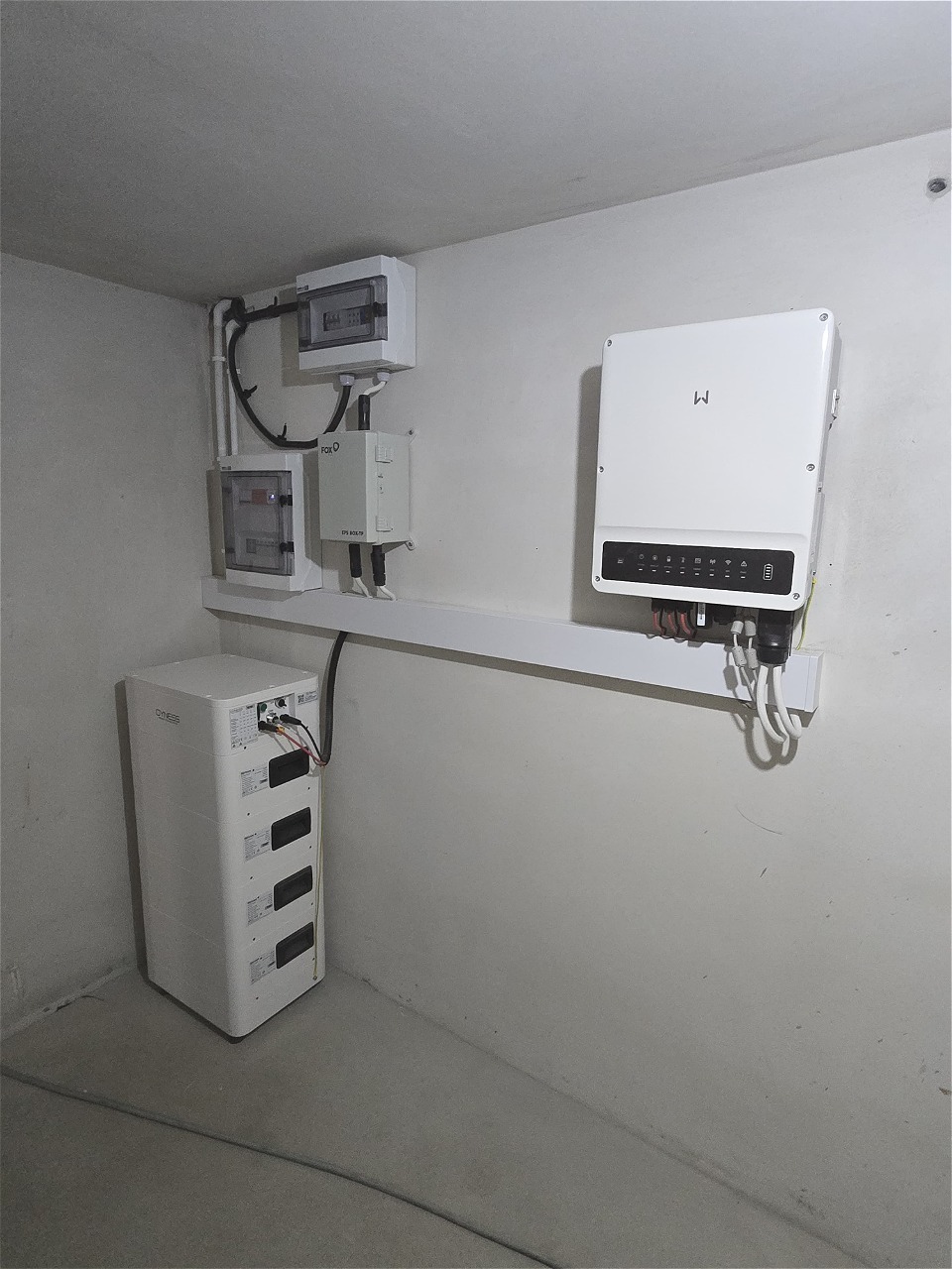 Biały magazyn energii z rozdzielnicą i inwerterem na ścianie w pomieszczeniu. Widoczne kable i przewody. Minimalistyczny design.