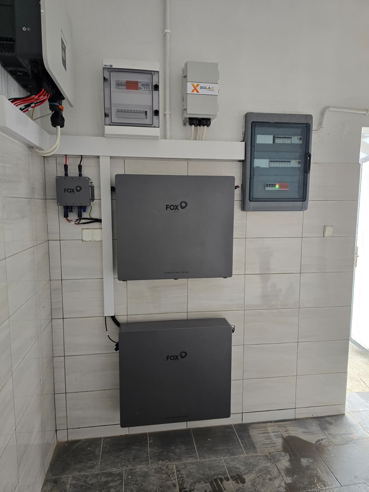 Dwa akumulatory FOX ESS zamontowane na kafelkowanej ścianie, obok skrzynki elektrycznej i inwertera Solax. Widoczne przewody i elementy instalacji fotowoltaicznej.