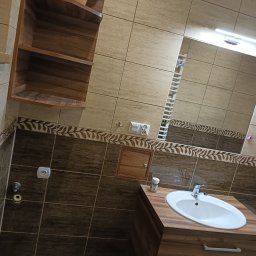 Pat bud - Ukośne ujęcie wykończonej łazienki z brązowo-beżowymi płytkami, drewnianą szafką pod umywalką, otwartą półką z kaktusem i dekoracyjnym pasem z motywem liści.