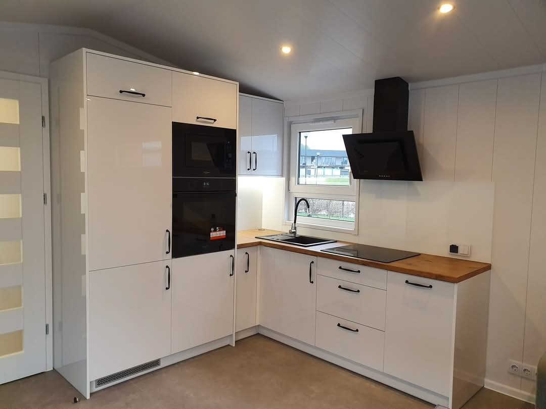 Nowoczesna kuchnia z białymi, lakierowanymi frontami, czarnymi uchwytami i drewnianym blatem, minimalistyczny okap nad płytą indukcyjną, widok z niskiego kąta.