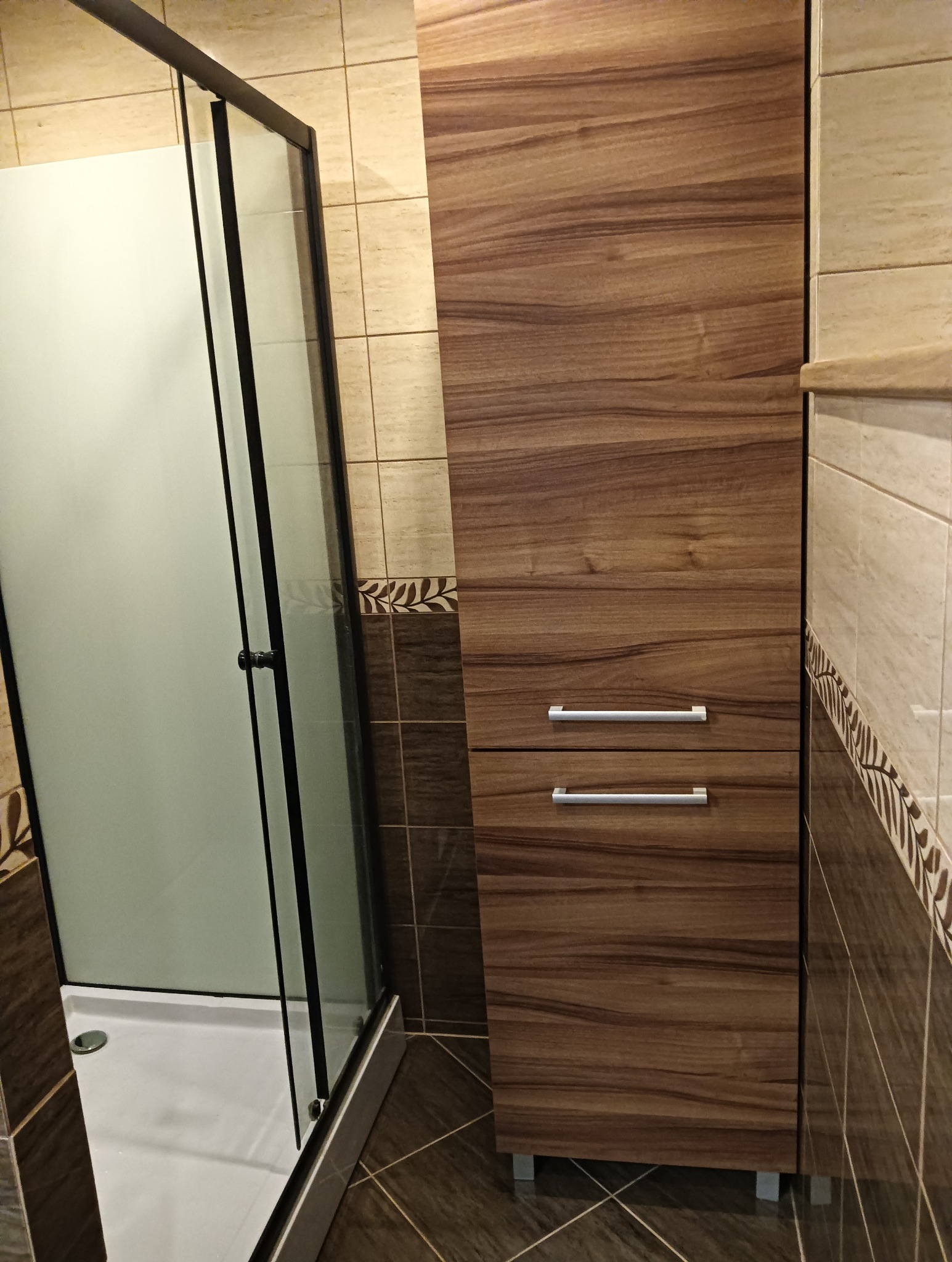 Nowoczesna kabina prysznicowa z matowego szkła i wysoka szafka łazienkowa w odcieniach brązu, dopasowane do płytek z motywem liści na ścianach.